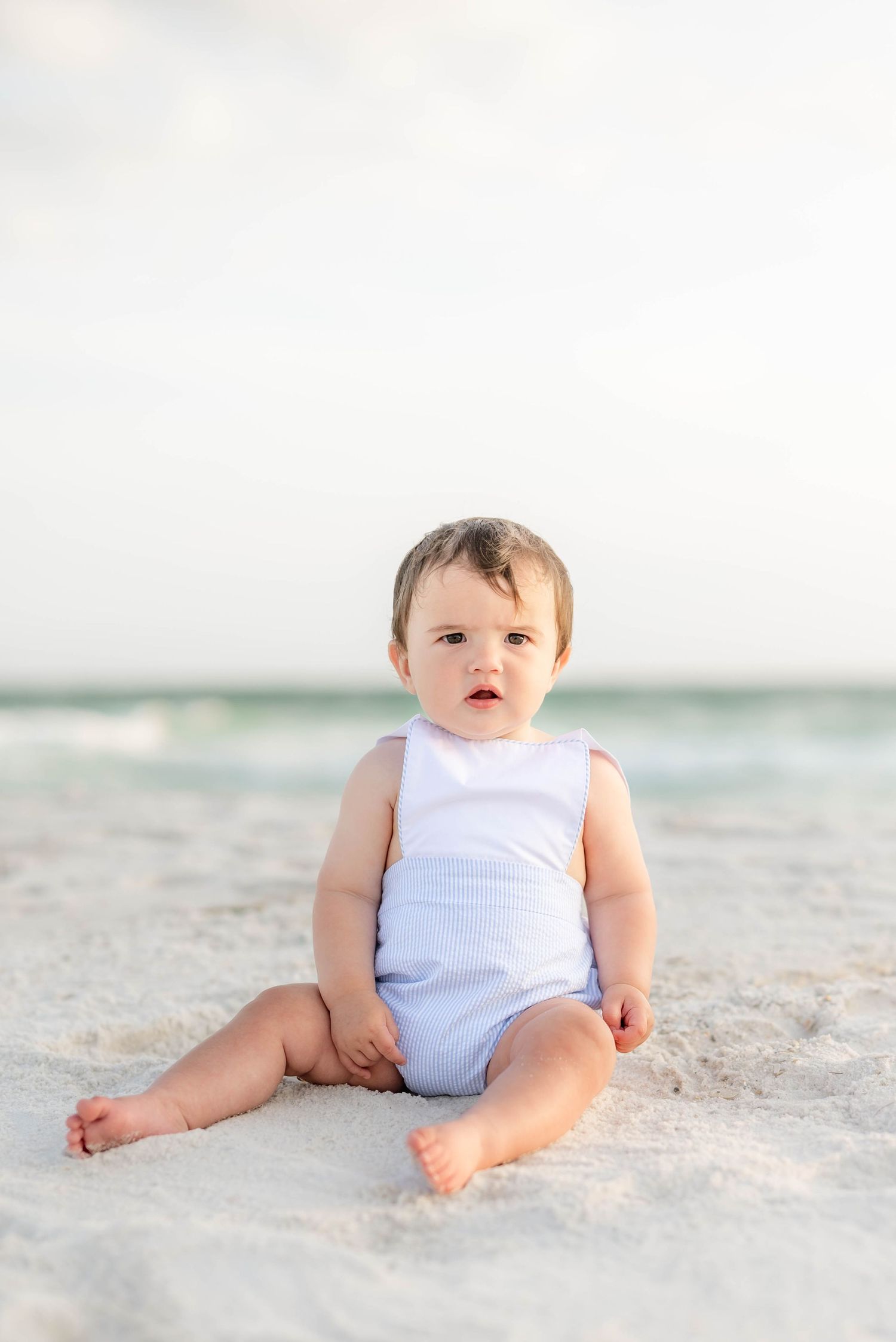 Baby in white outfit sitting on pristine sandy beach with ocean waves in background in Panama City Beach, Fl..