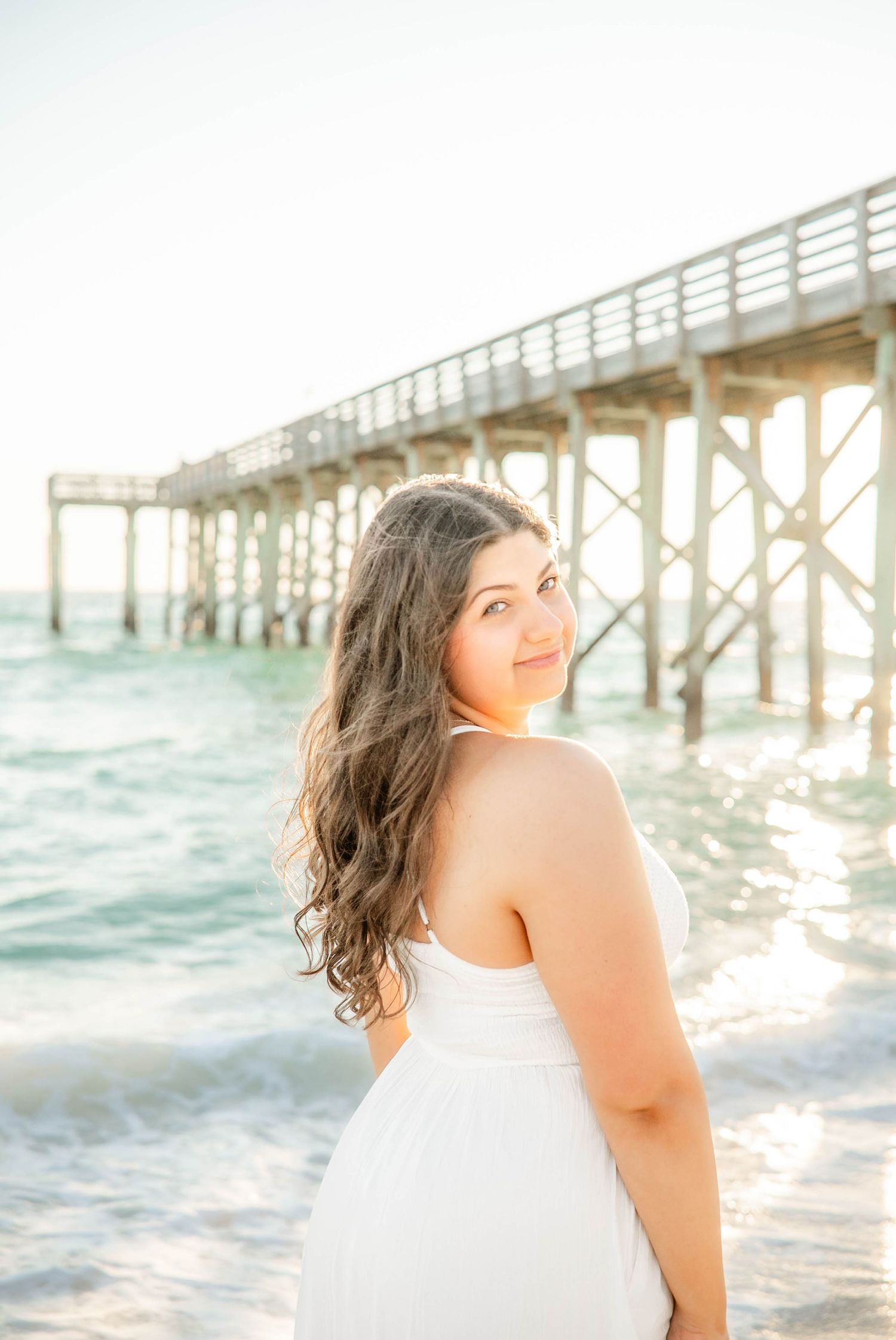 Woman in white dress poses near a wooden pier at the beach during golden hour.