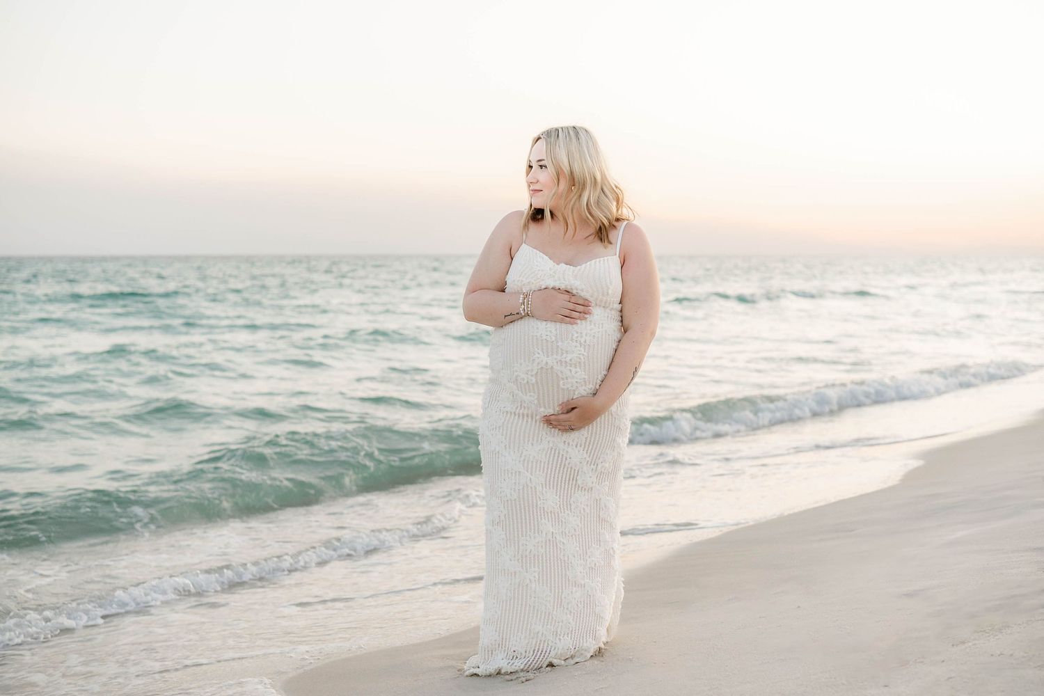 Expectant mother in flowing white dress walking along ocean shore at sunset in Panama City Beach, Fl..