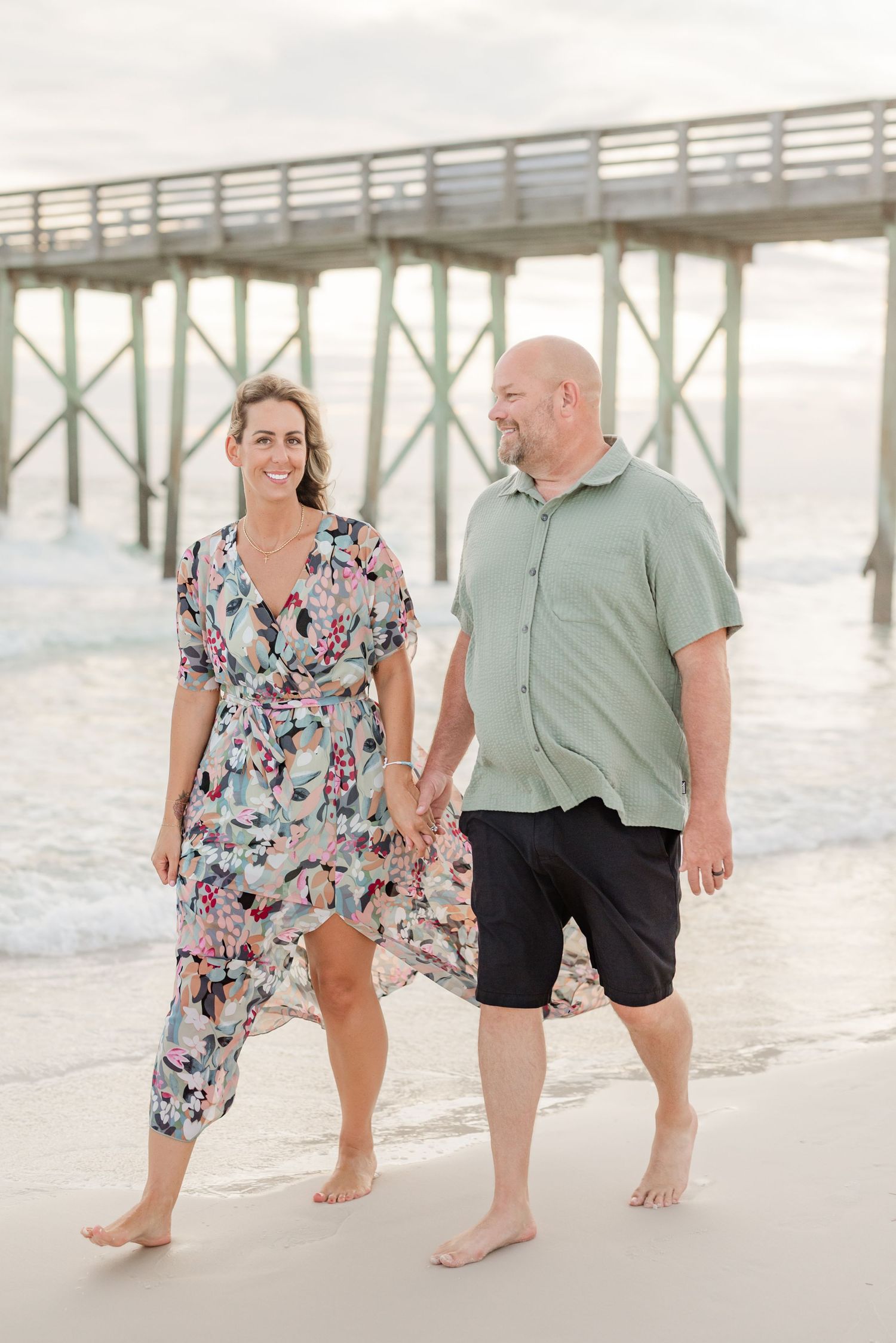 Romantic couple strolling barefoot on beach near wooden pier during golden hour.