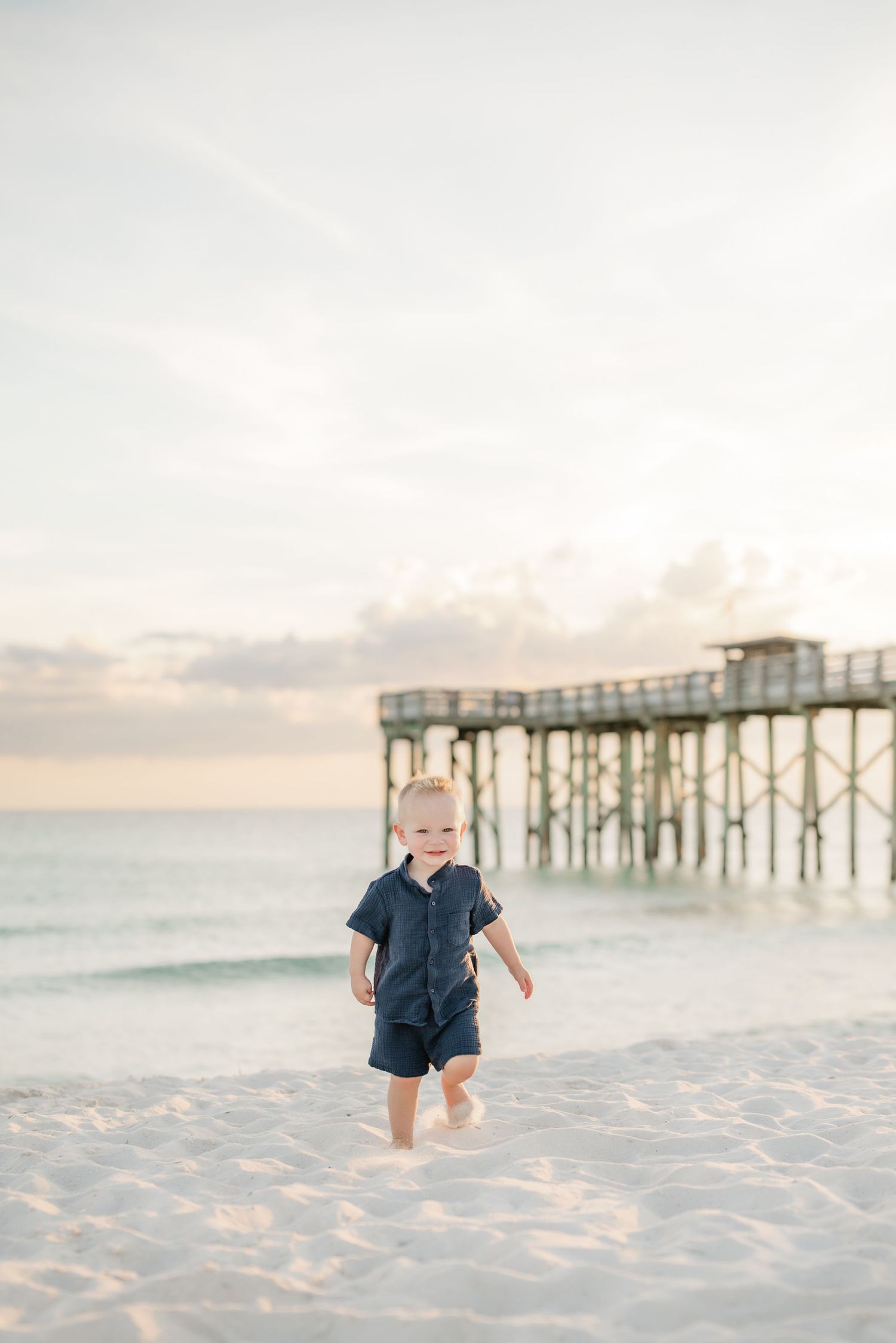 A child in navy blue outfit plays on beach sand with wooden pier in sunset background.