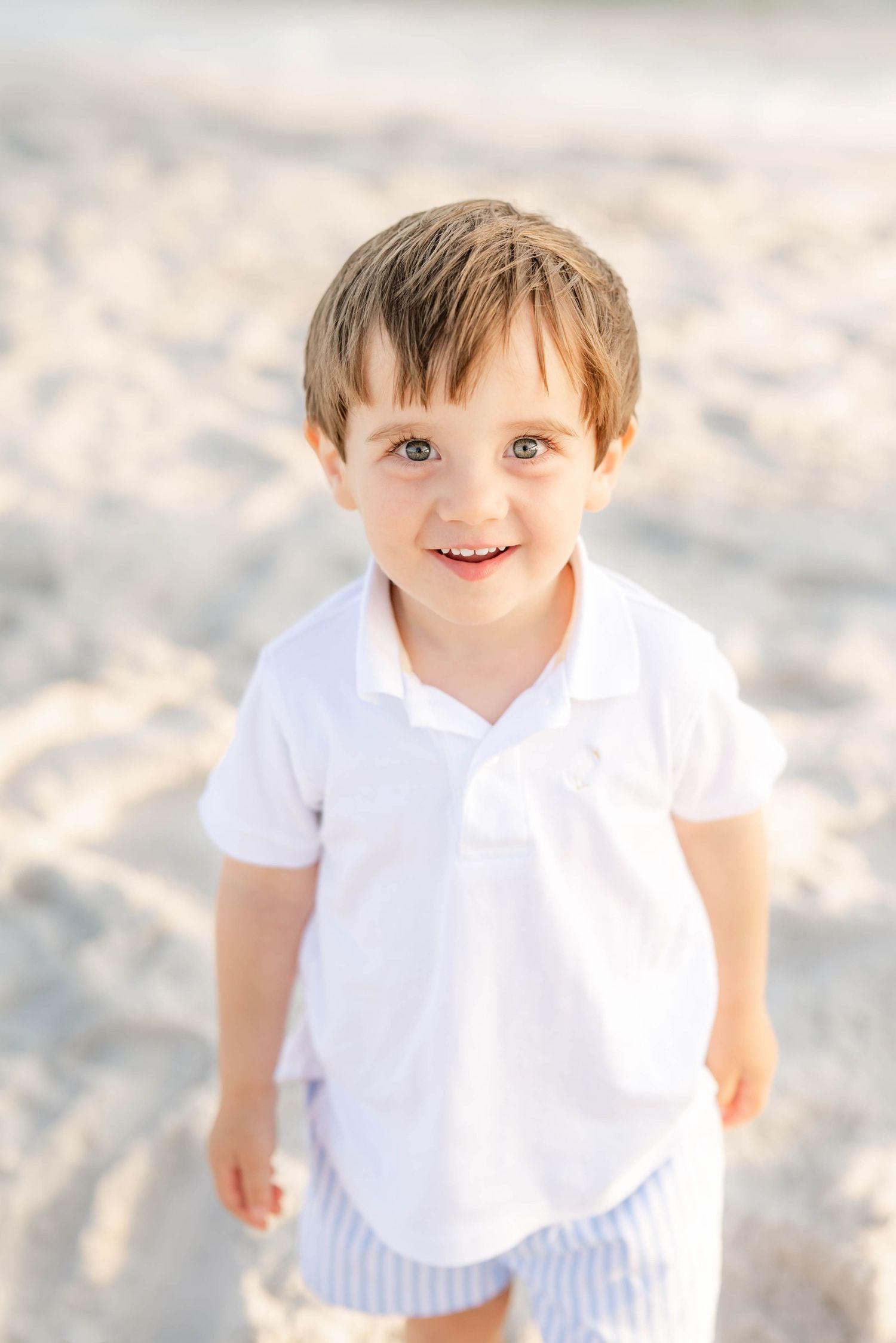 Child wearing white polo and striped shorts playing on sunny beach.
