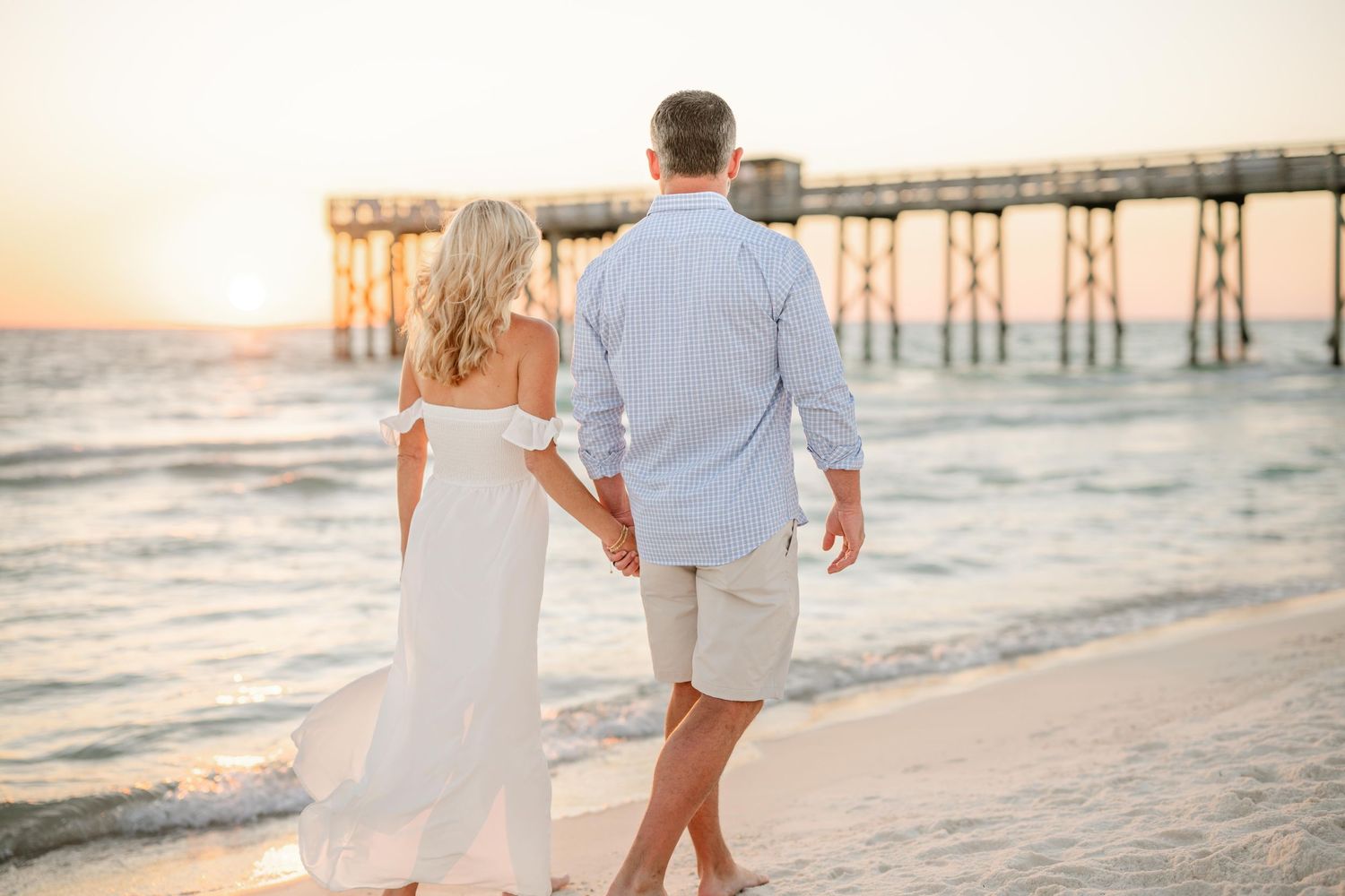 A couple walks hand in hand along the beach near a wooden pier at sunset.
