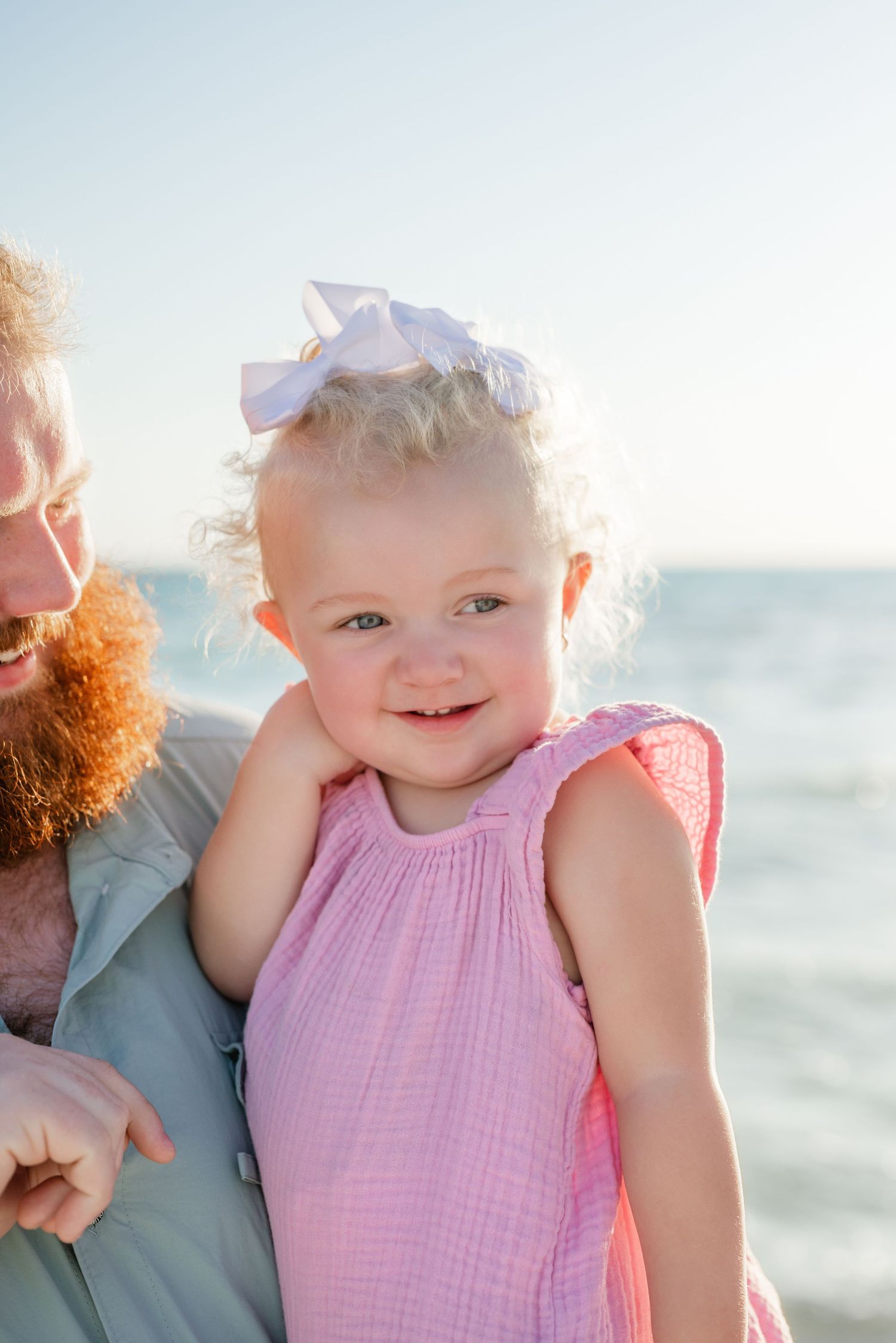 A smiling toddler in a pink dress poses on the beach at sunset.