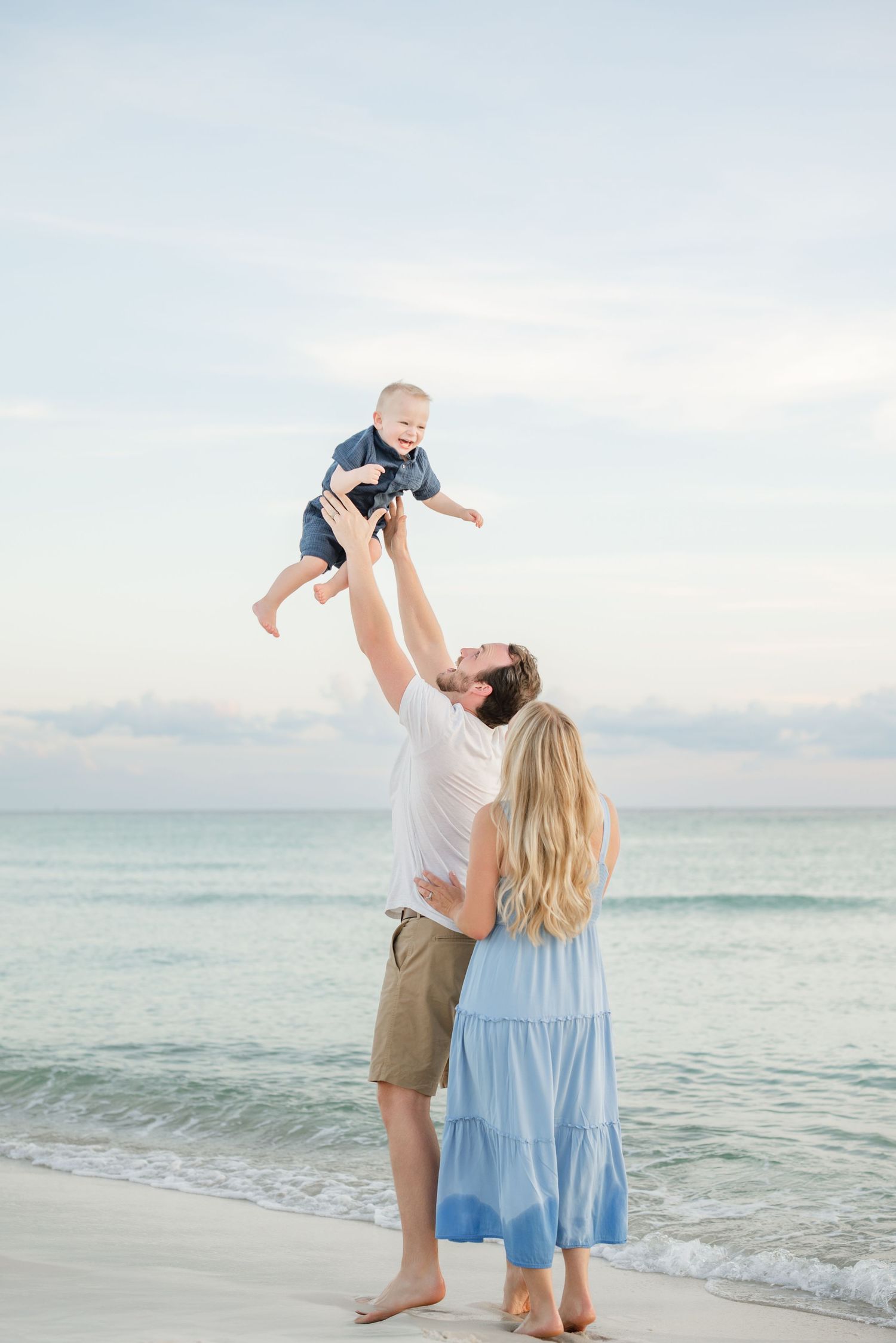 A family plays and lifts their child in the air while enjoying a beach sunset.
