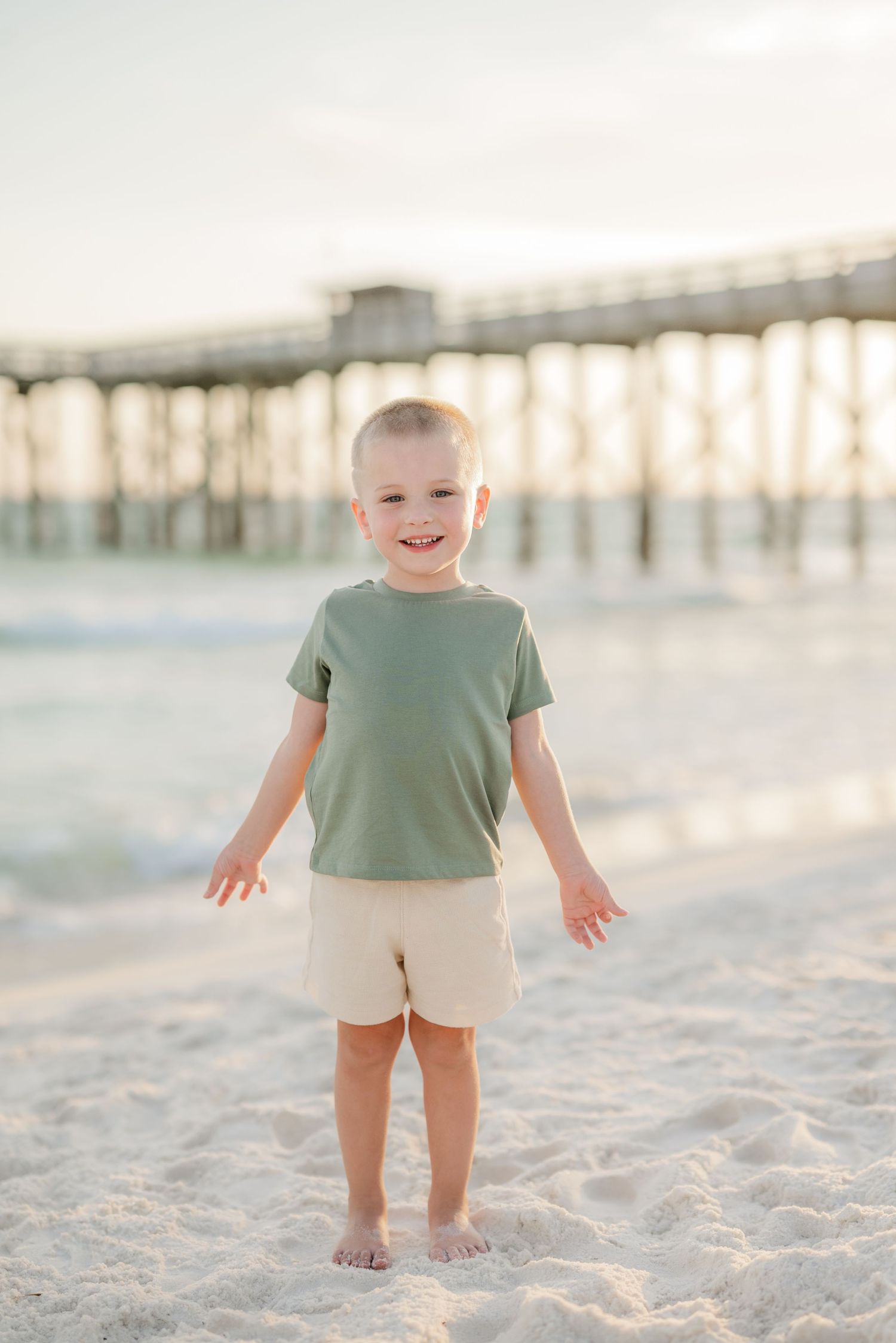 A child in a sage green shirt and beige shorts stands joyfully on a sandy beach with a pier in the background.