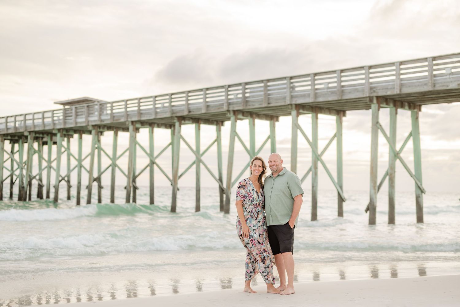 A wooden pier stretches into the ocean under cloudy skies at the beach.