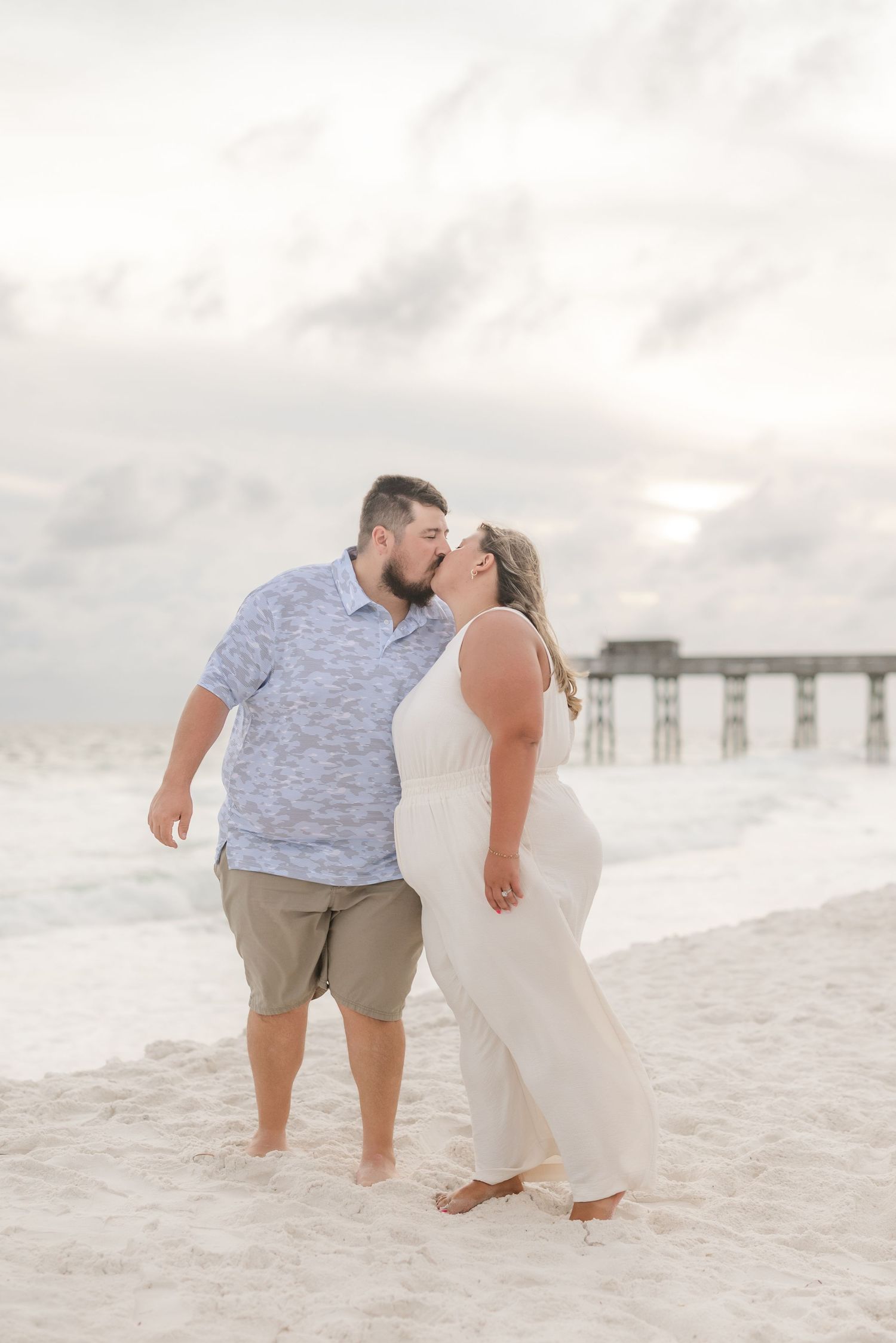 Couple in light summer clothing embracing on pristine beach with pier in background.
