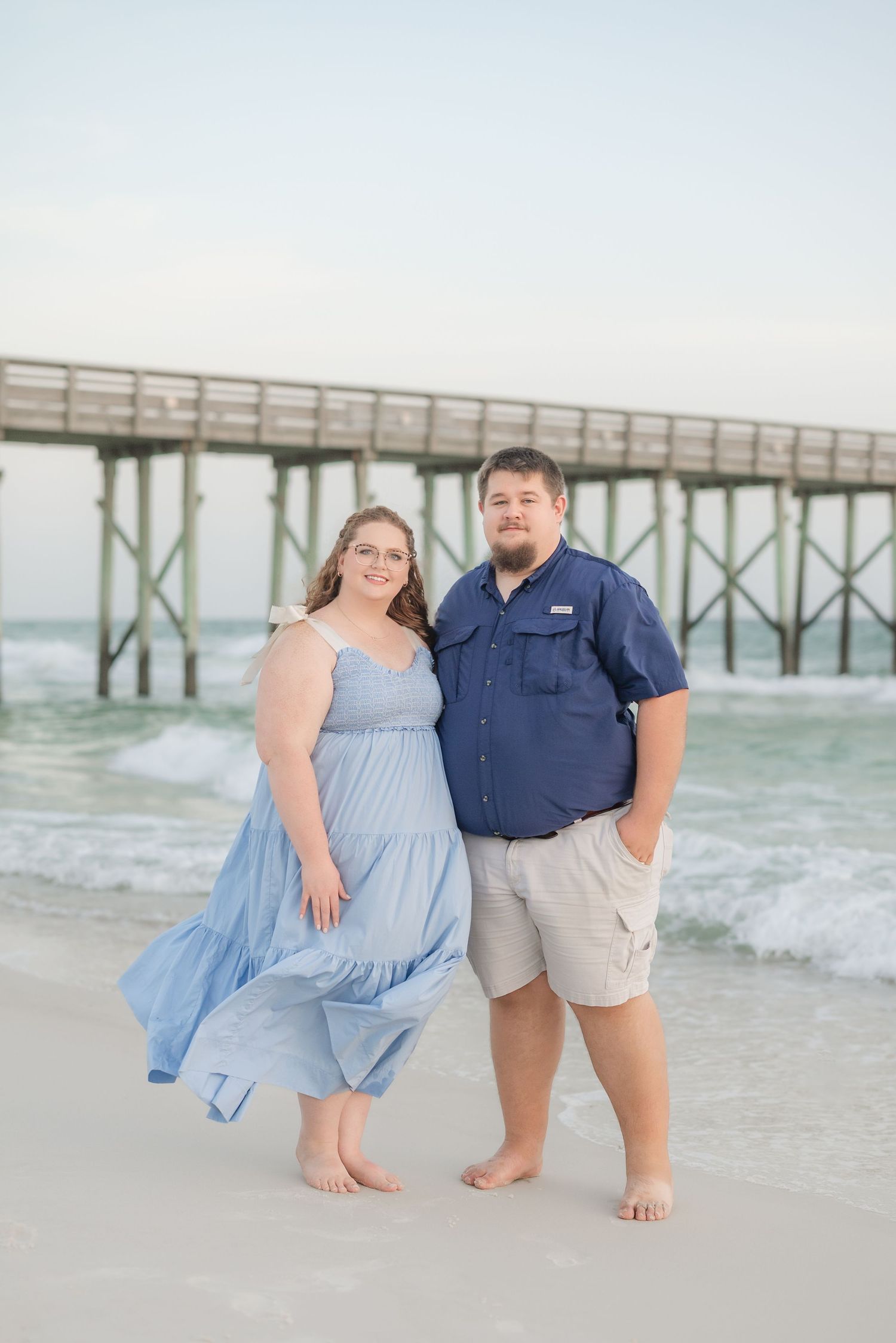 Couple poses casually by wooden pier in beachwear during overcast day.