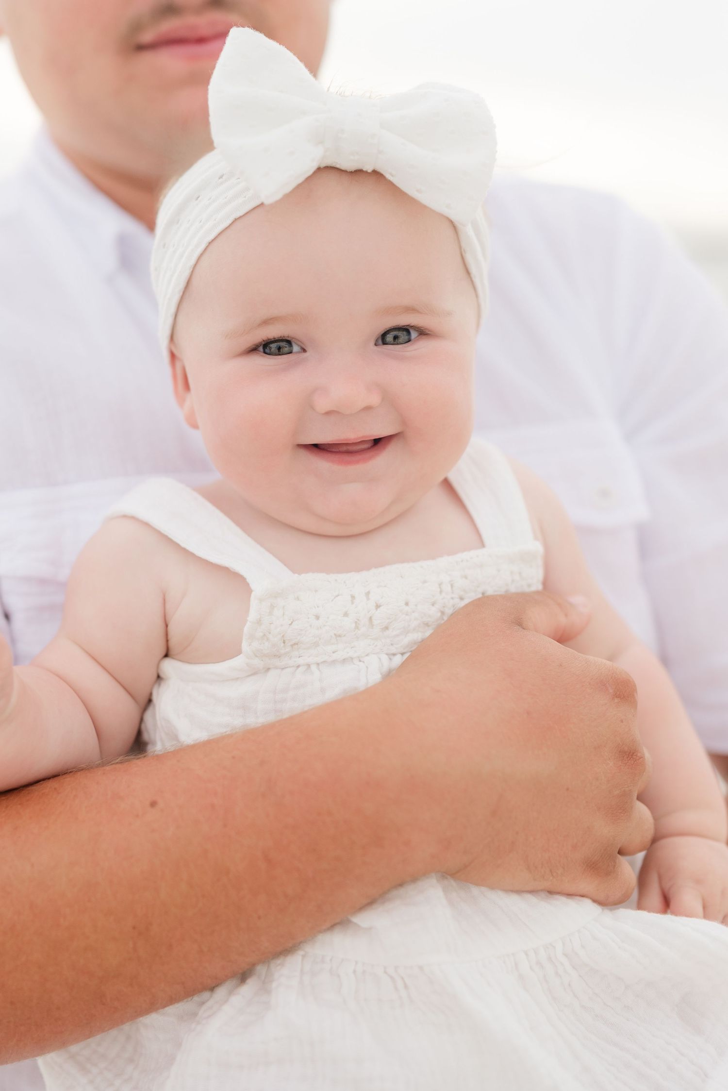 Baby wearing white bow headband and dress smiles while being held in Panama City Beach, Fl..