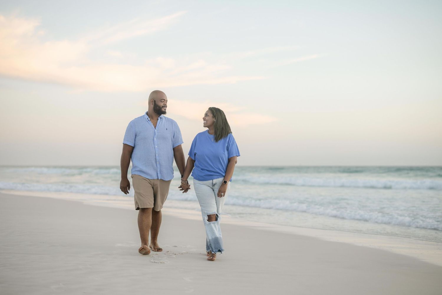 Couple holding hands walking on a sandy beach at sunset with ocean waves in the background.