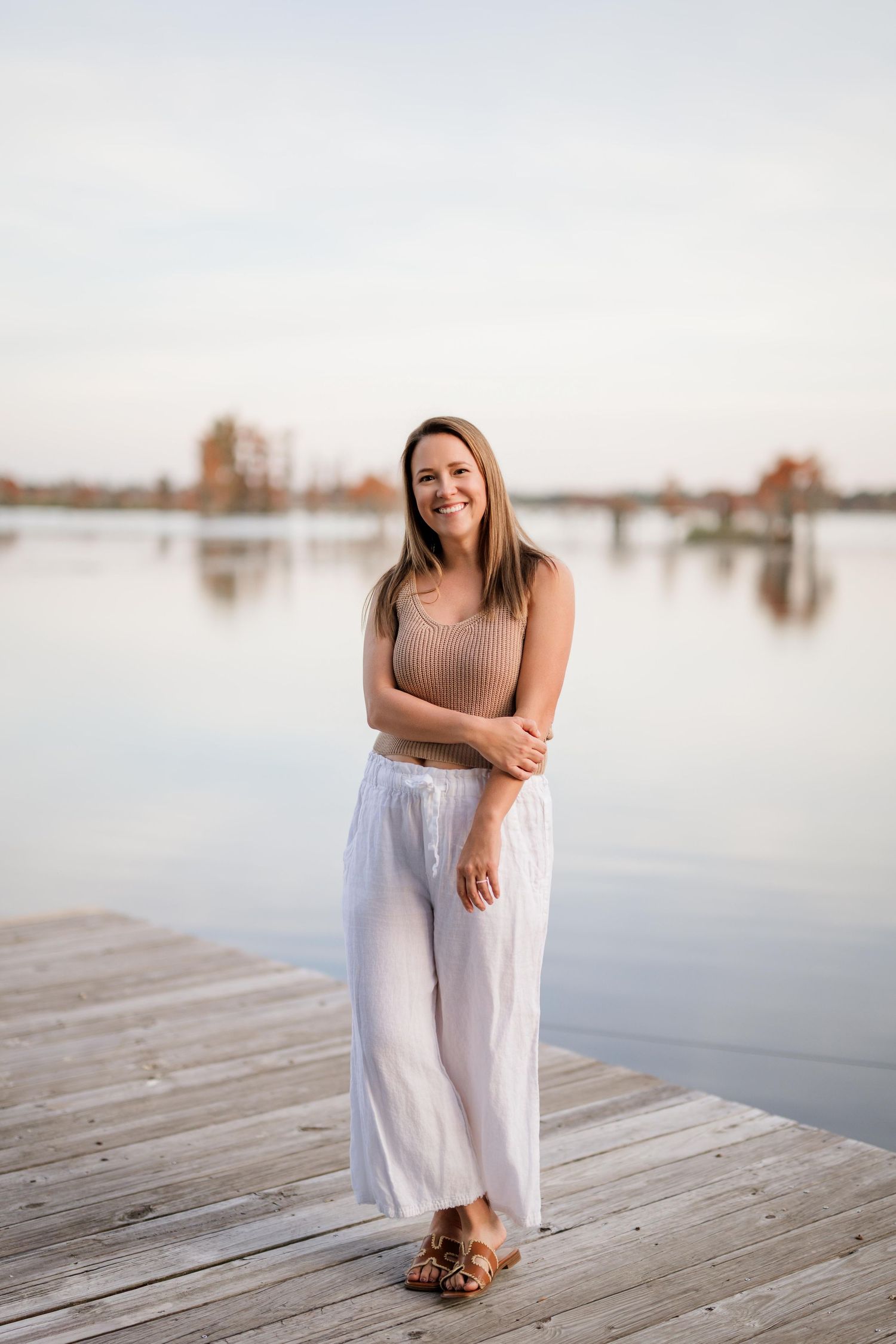 Woman in white dress sitting on wooden dock by tranquil water during sunset photoshoot.
