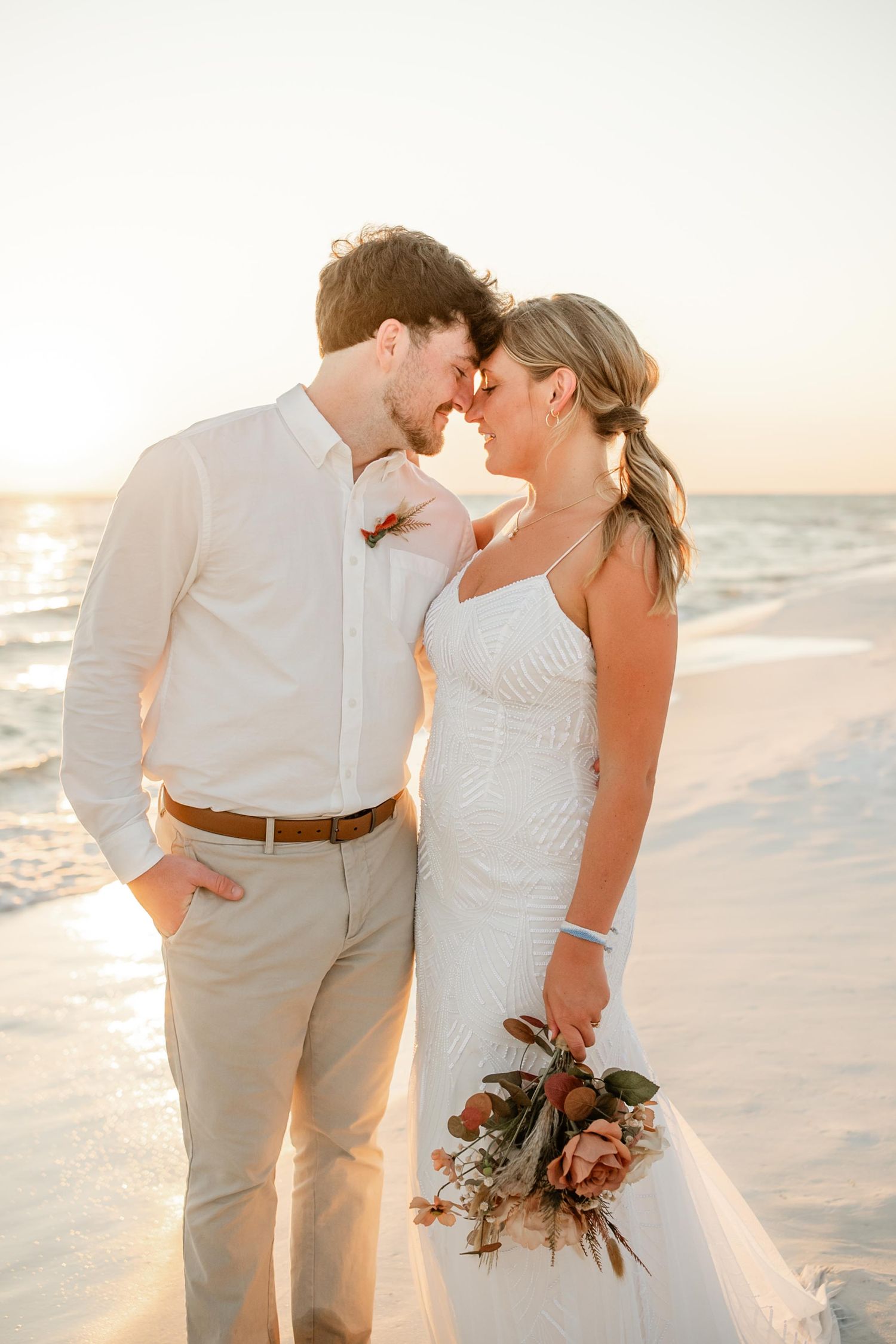Romantic beach wedding couple embraces at sunset with soft golden light and floral bouquet.