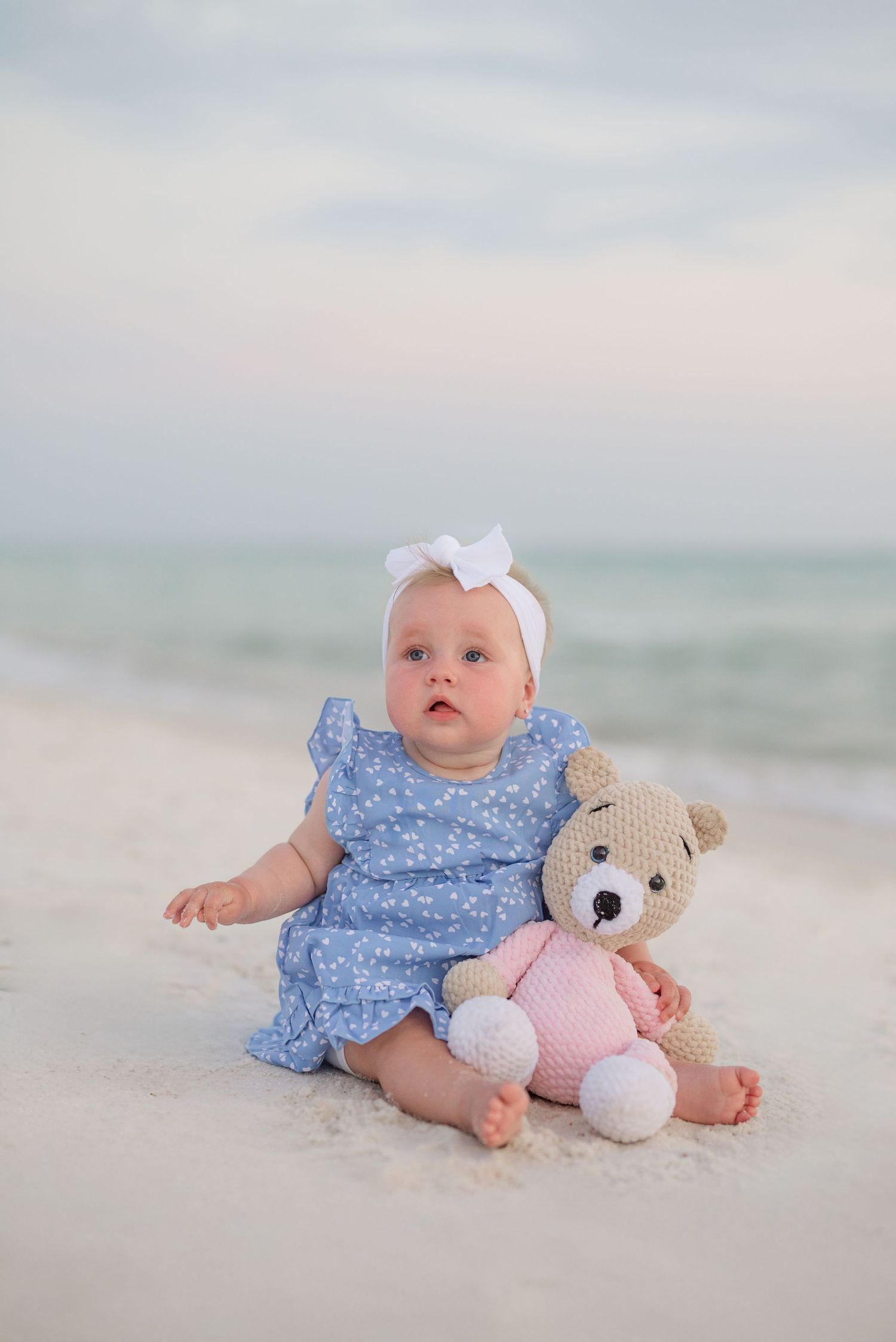 A baby in a blue dress sits on beach sand with a plush teddy bear toy.