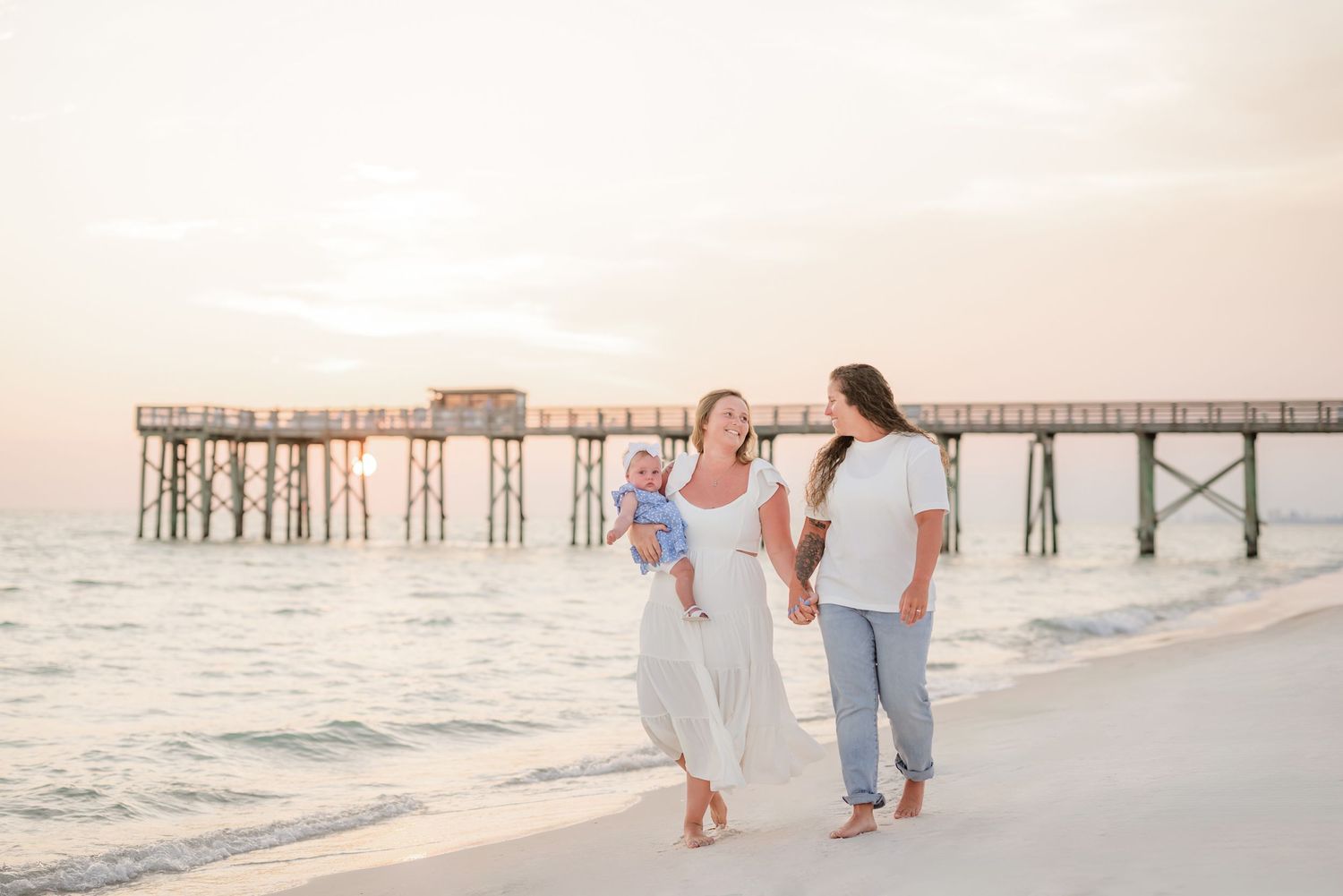 A family walks along a beach at sunset with a wooden pier in the background.