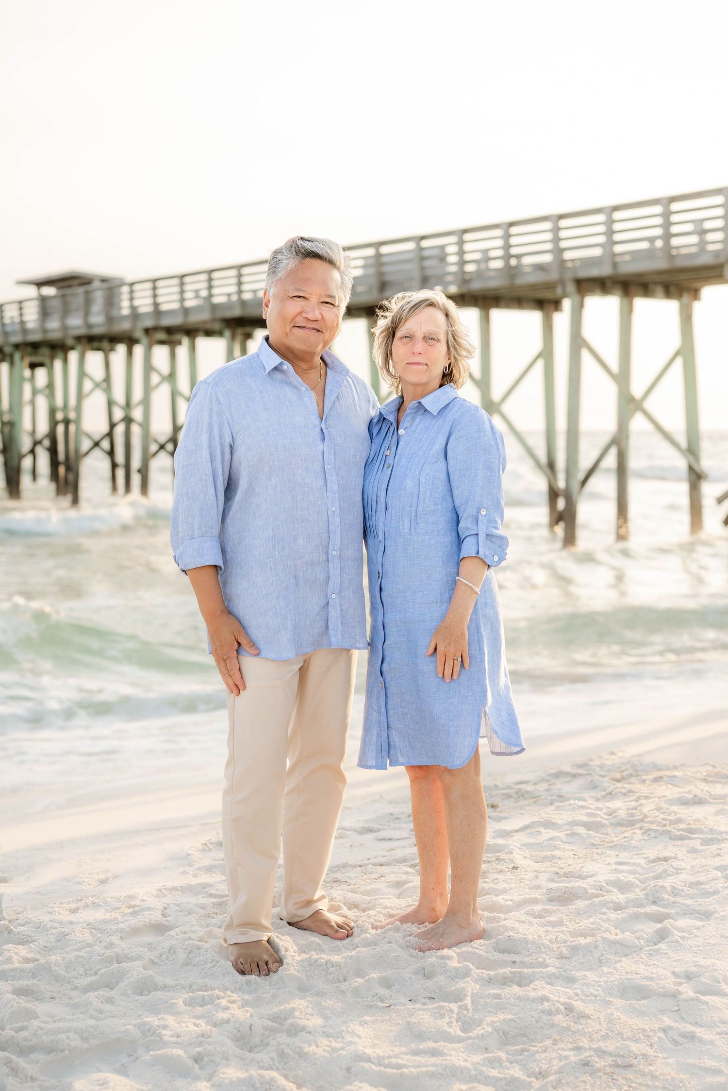 A couple in matching blue outfits poses for a beach portrait near a wooden pier.