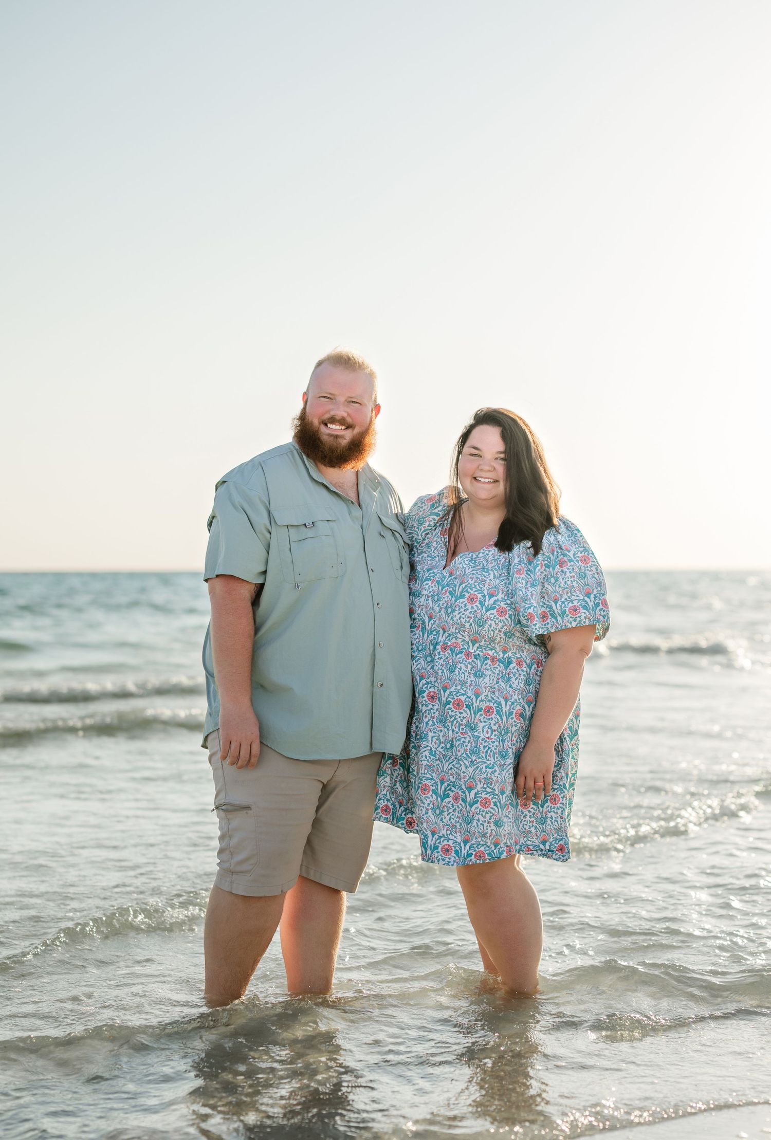A couple stands together in the ocean waves during a beach photoshoot at sunset.
