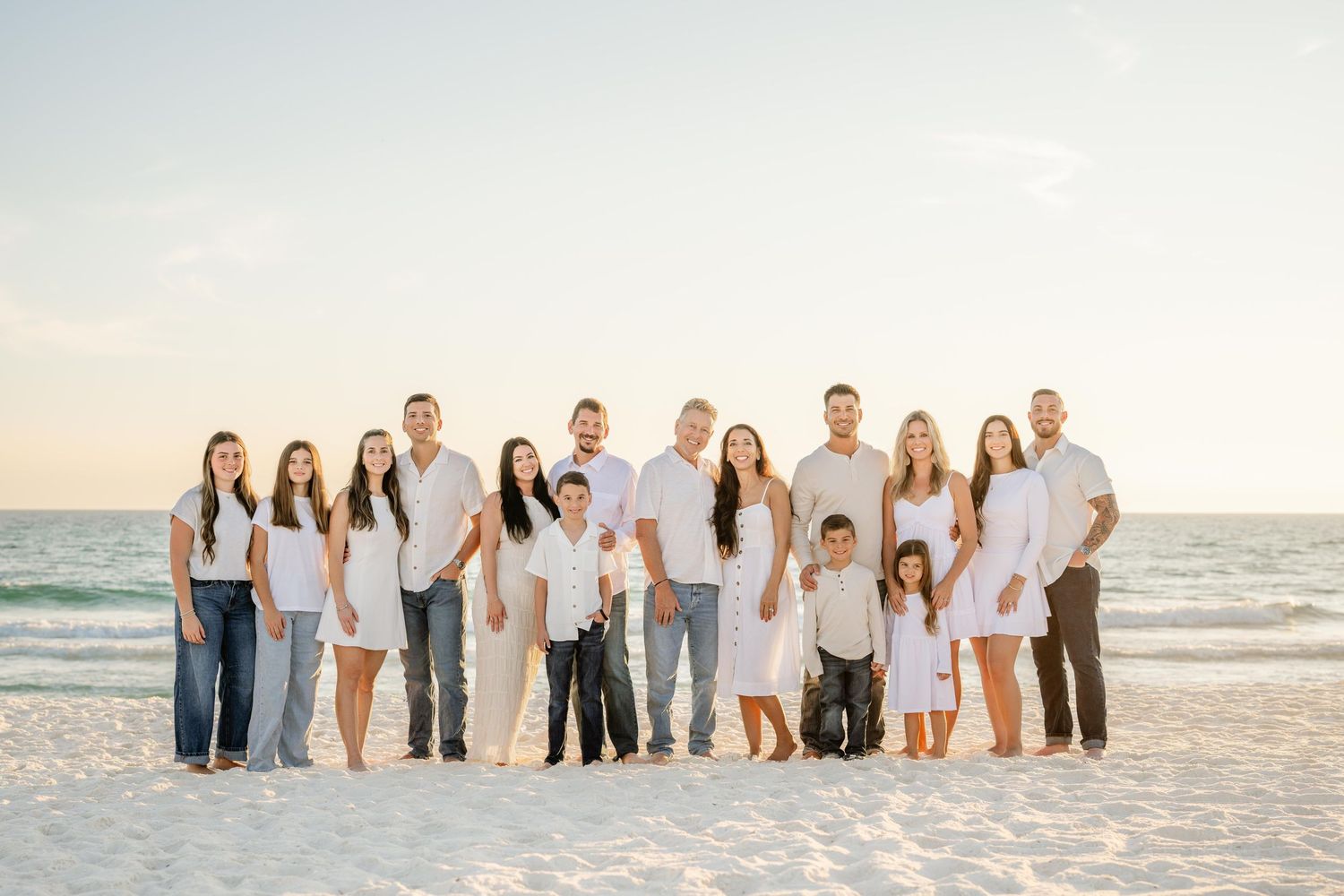 Large family group wearing white and neutral colors poses together on sandy beach at sunset.