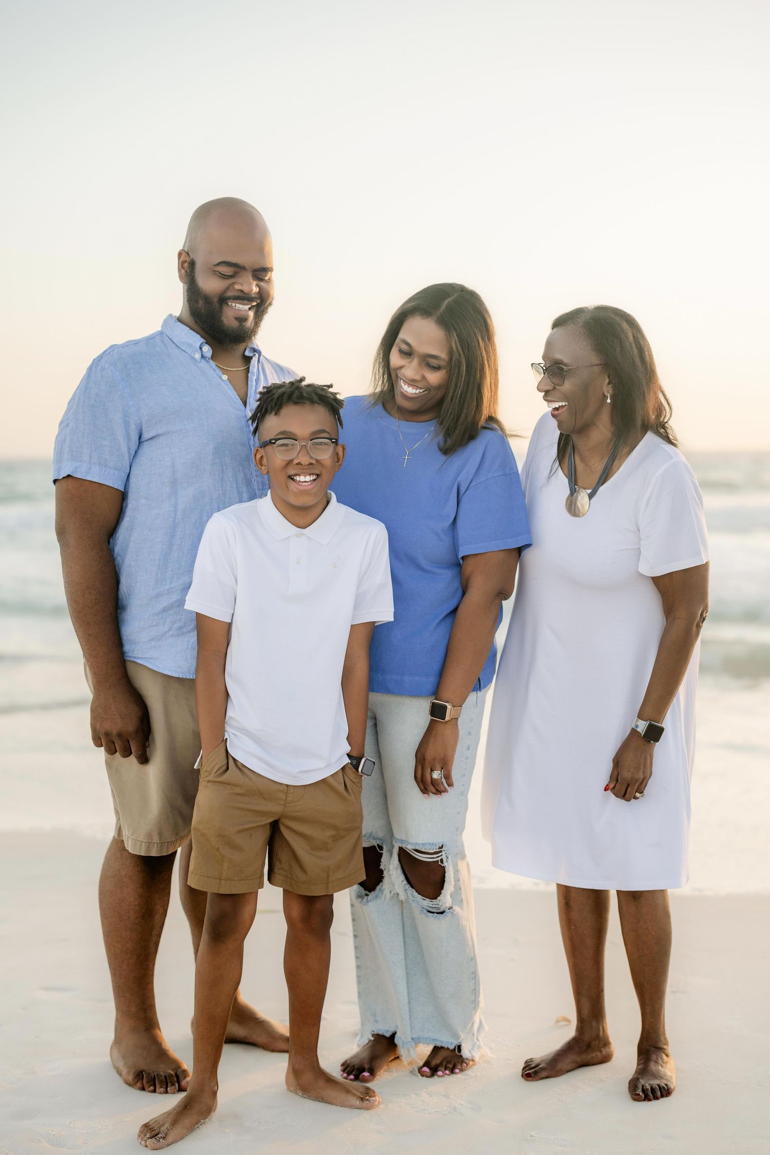 A joyful Black family of four poses barefoot on a white sand beach at sunset, smiling together in casual blue and white outfits.