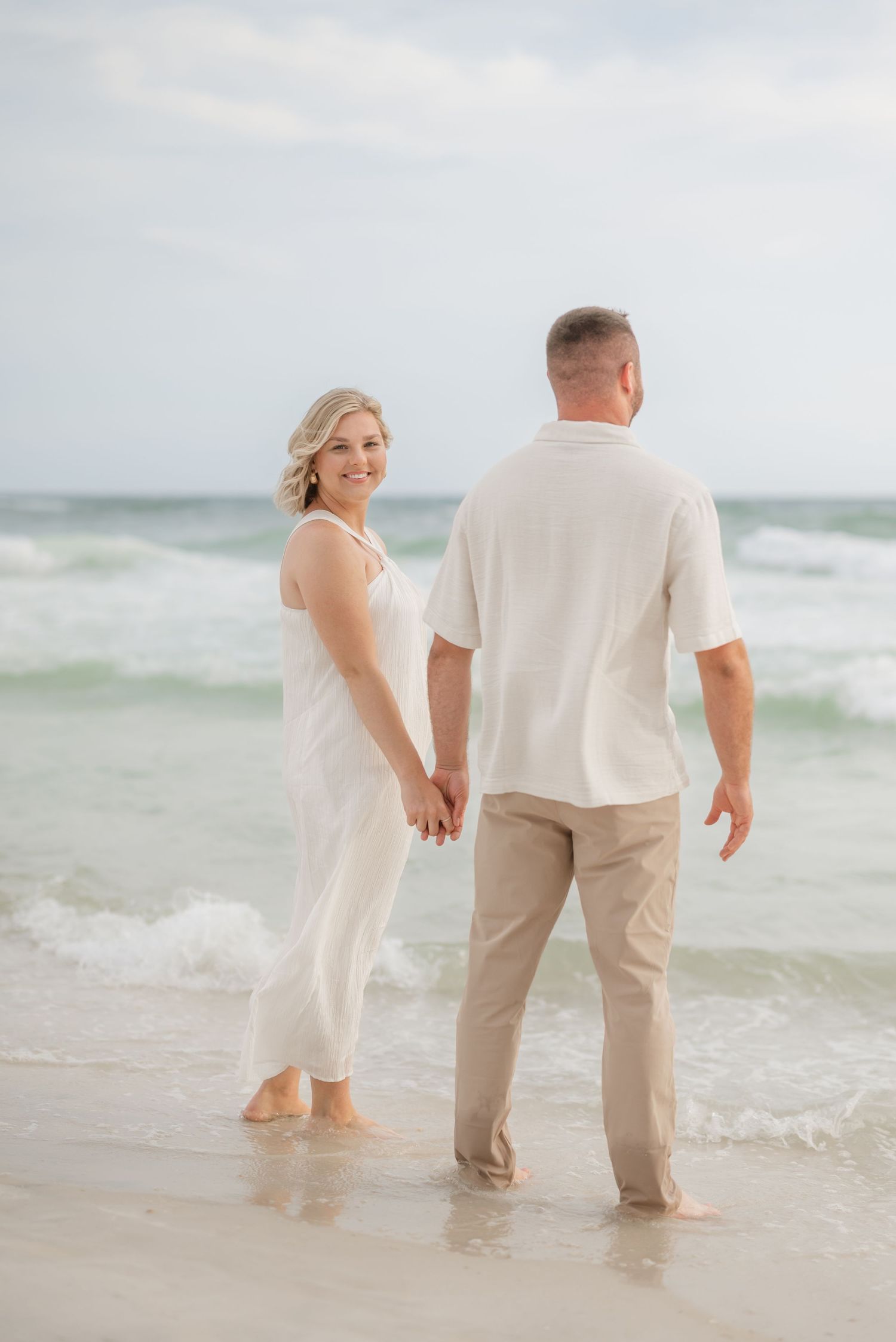 Couple wading in gentle ocean waves wearing coordinated white and beige outfits.