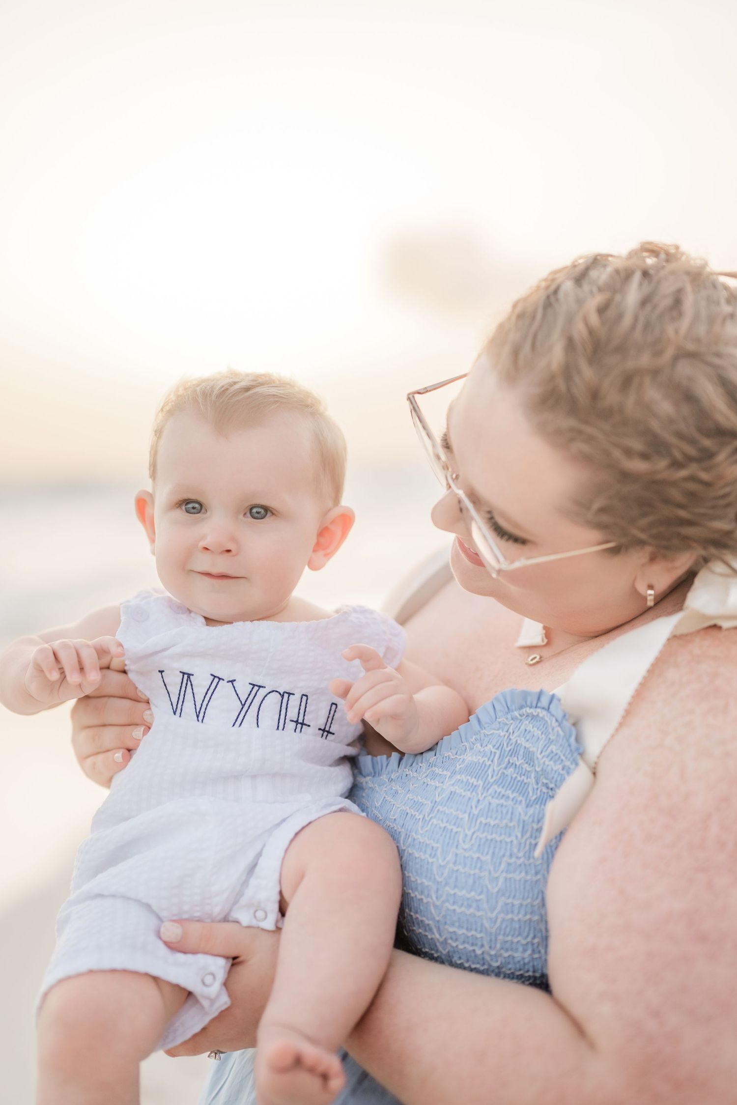 Baby in light blue outfit cuddling with parent during beach photoshoot.