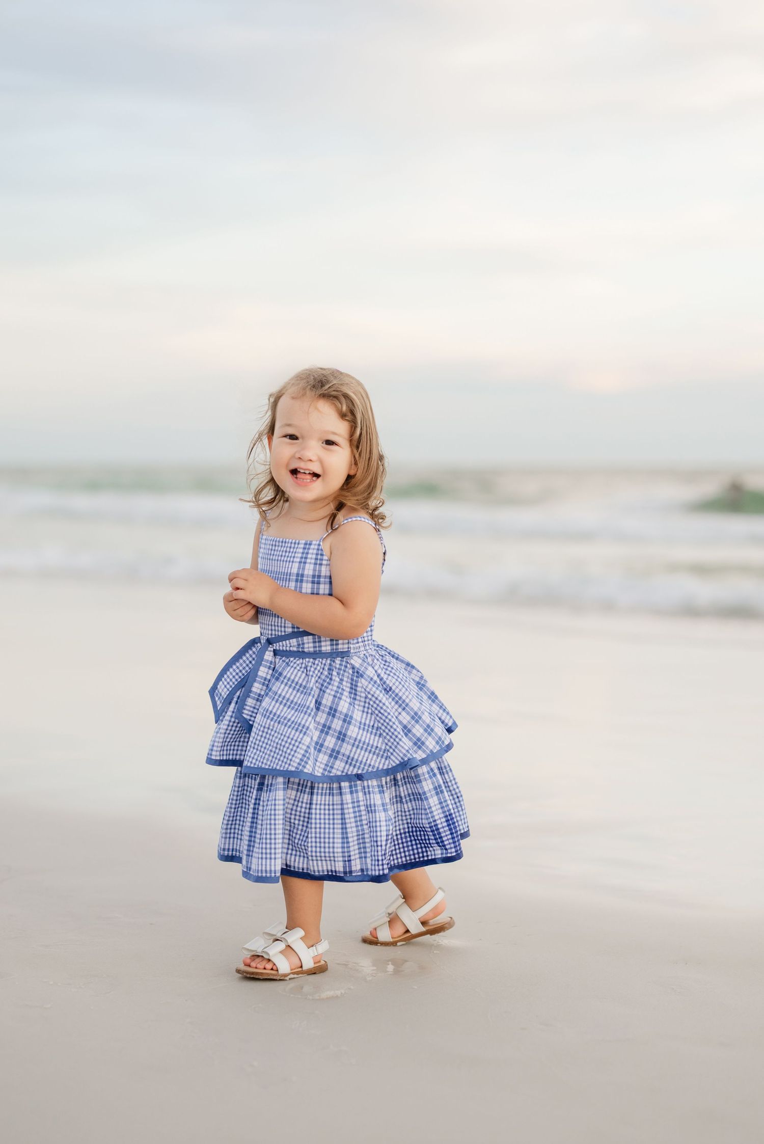 Child in blue gingham dress crouching playfully on sandy beach.