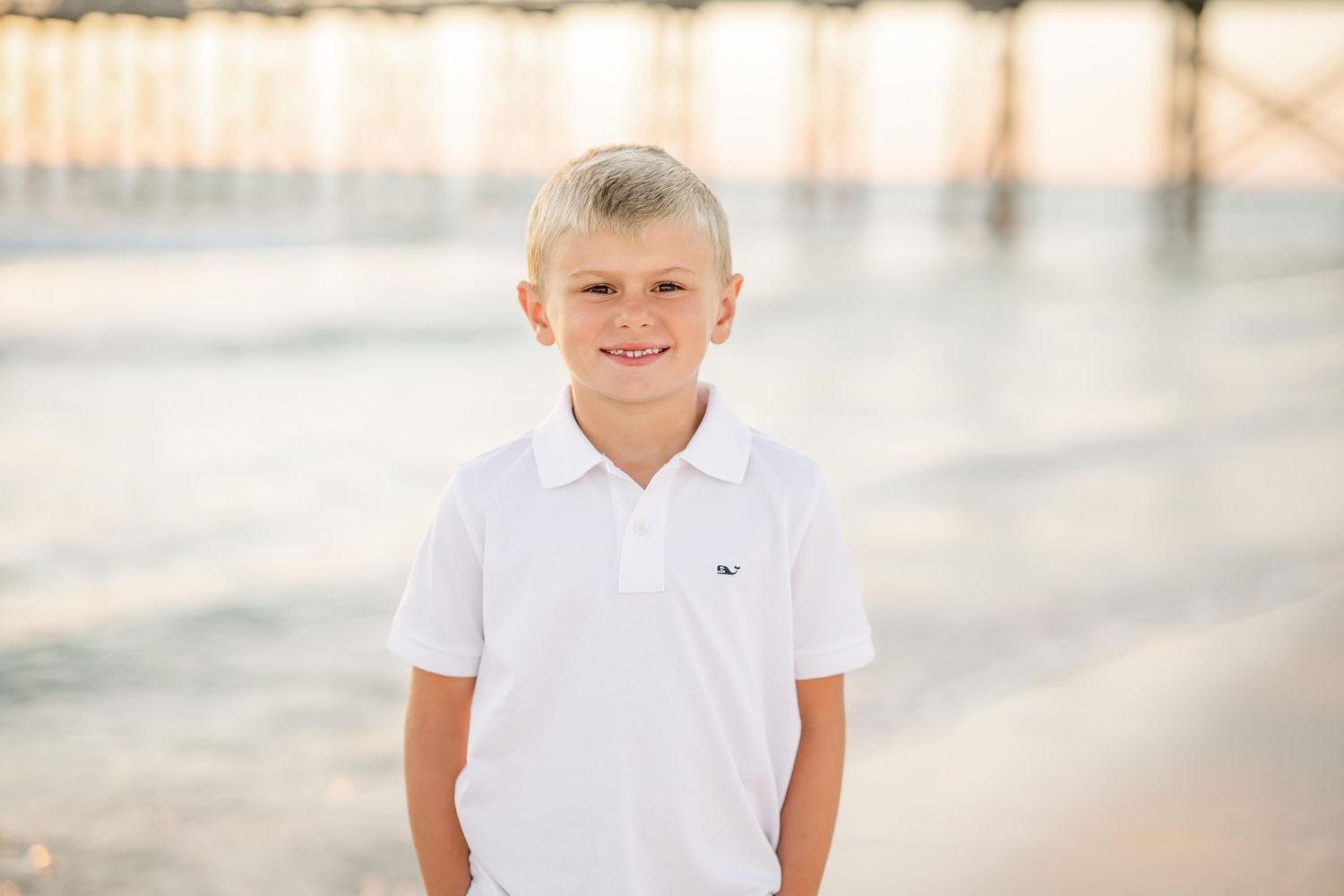 A person in white polo shirt stands on sandy beach with ocean pier in golden sunset light.