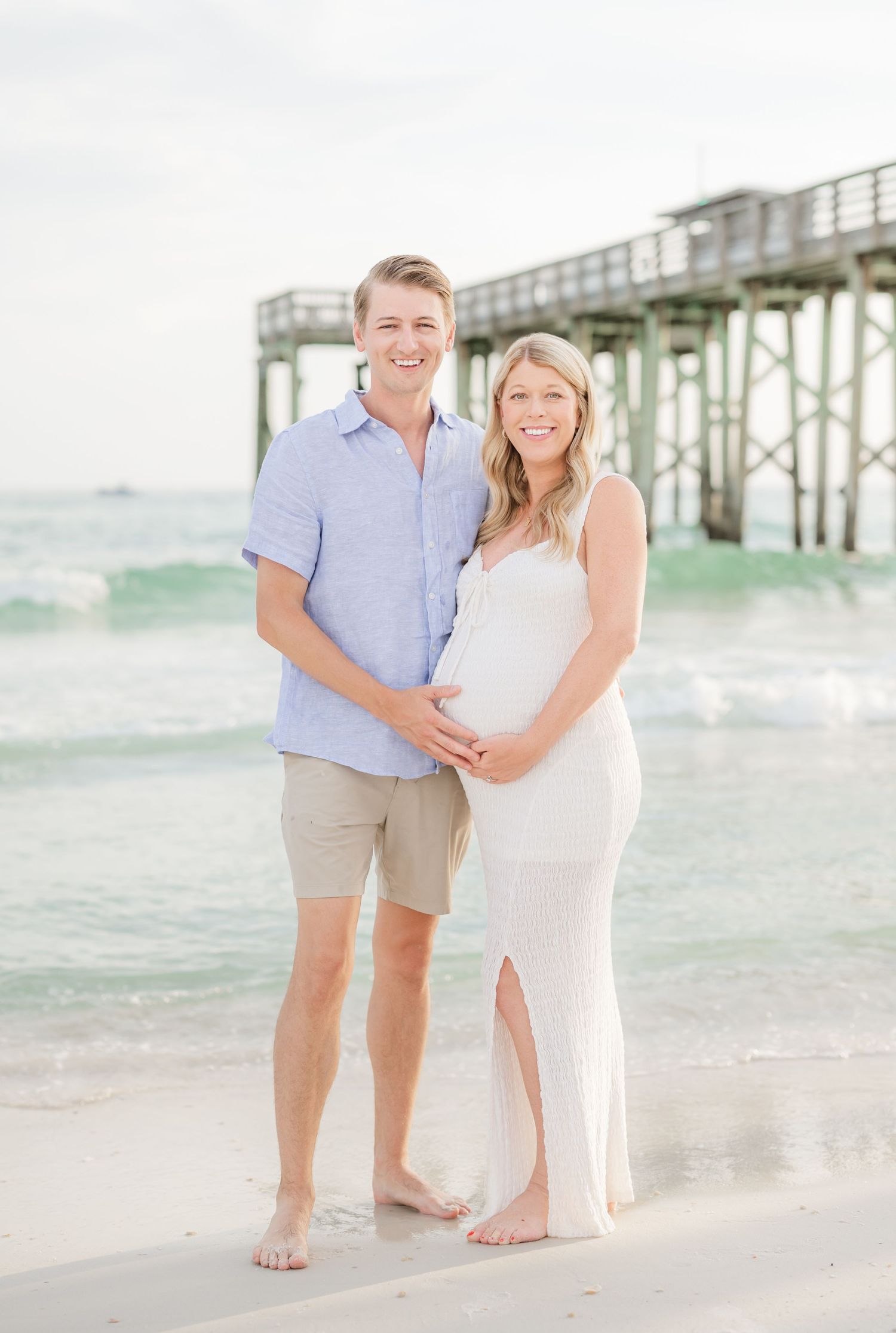 A couple poses for a maternity photo on the beach near a pier.