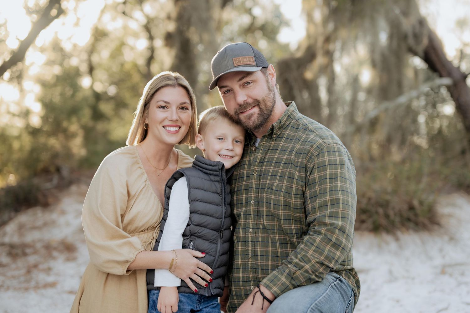 A family poses together outdoors in casual attire against a natural wooded background.