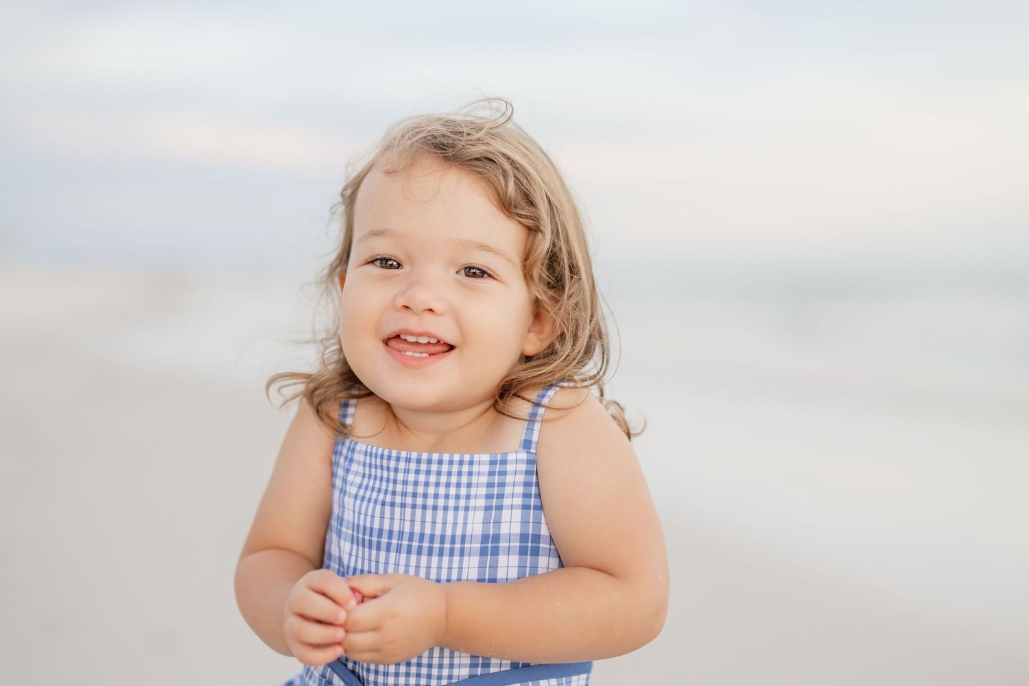 Small child in blue checkered dress poses playfully on beach at dusk.