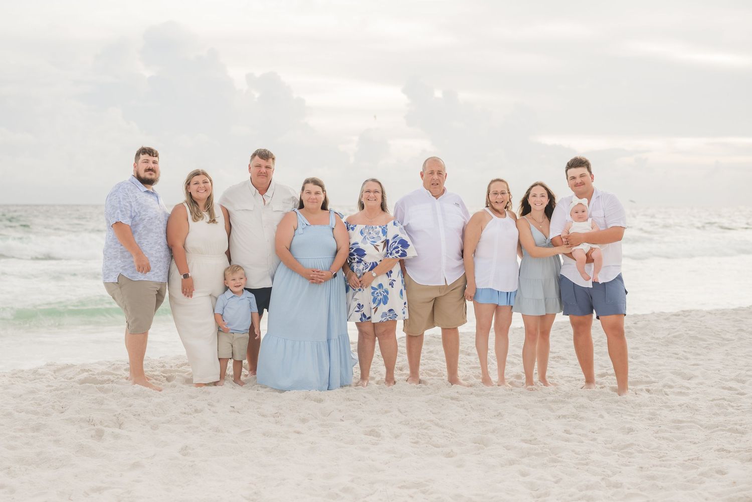 Large family group portrait on a sandy beach at sunset, wearing coordinated blue and white outfits.
