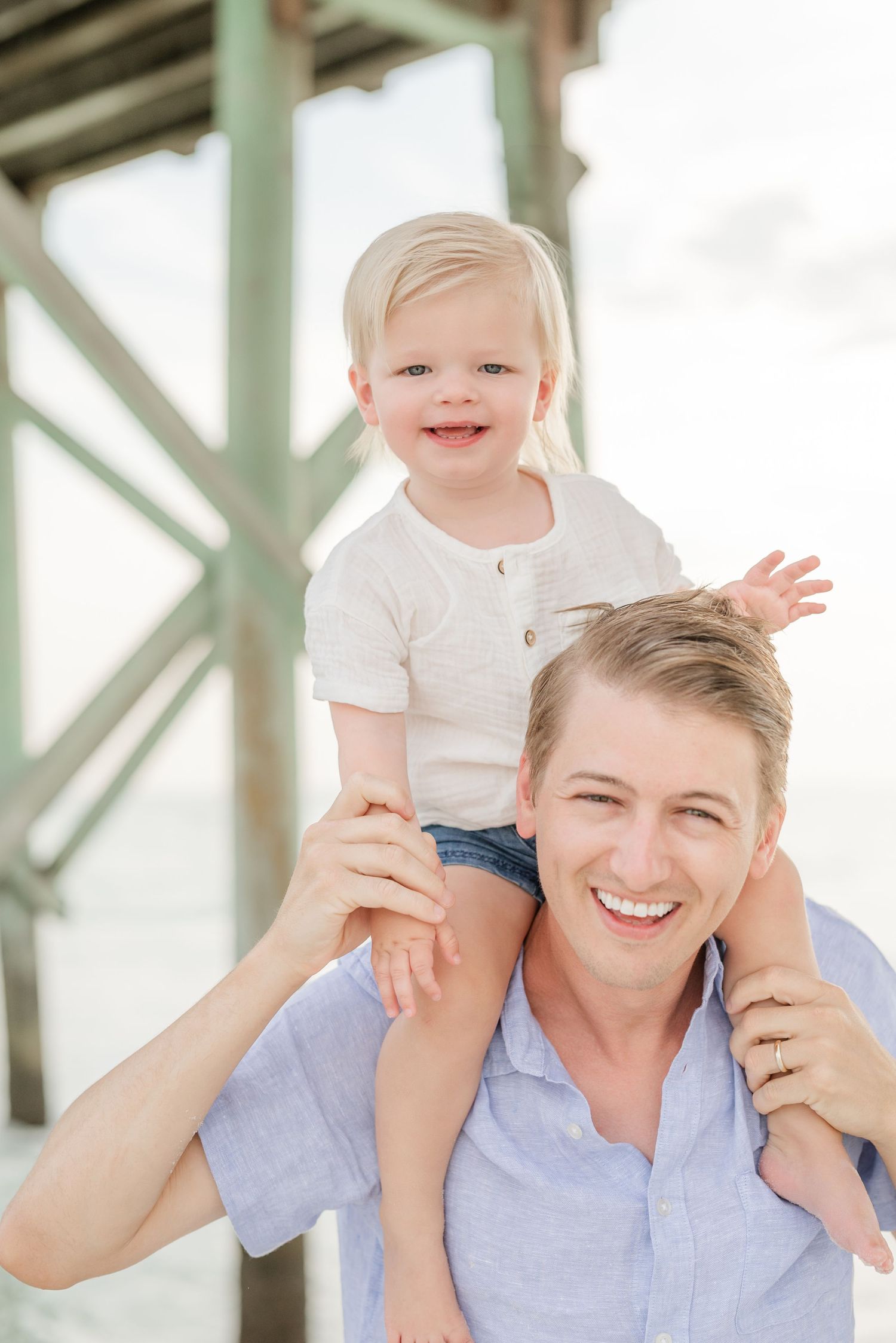 Smiling father carries toddler on shoulders at beach pier, both in light blue and white outfits.