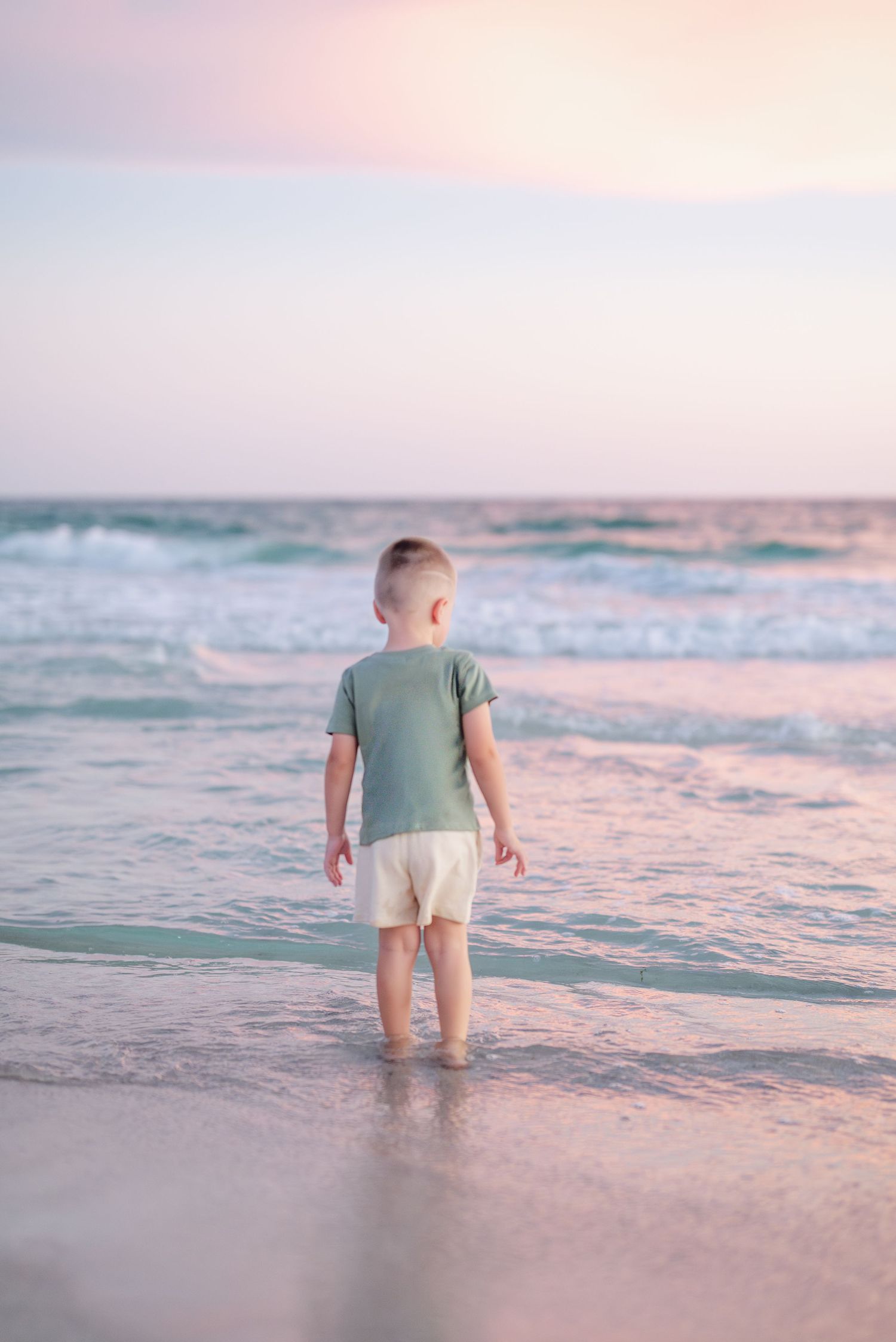 A child in casual beachwear stands at the ocean's edge during sunset with gentle waves approaching.