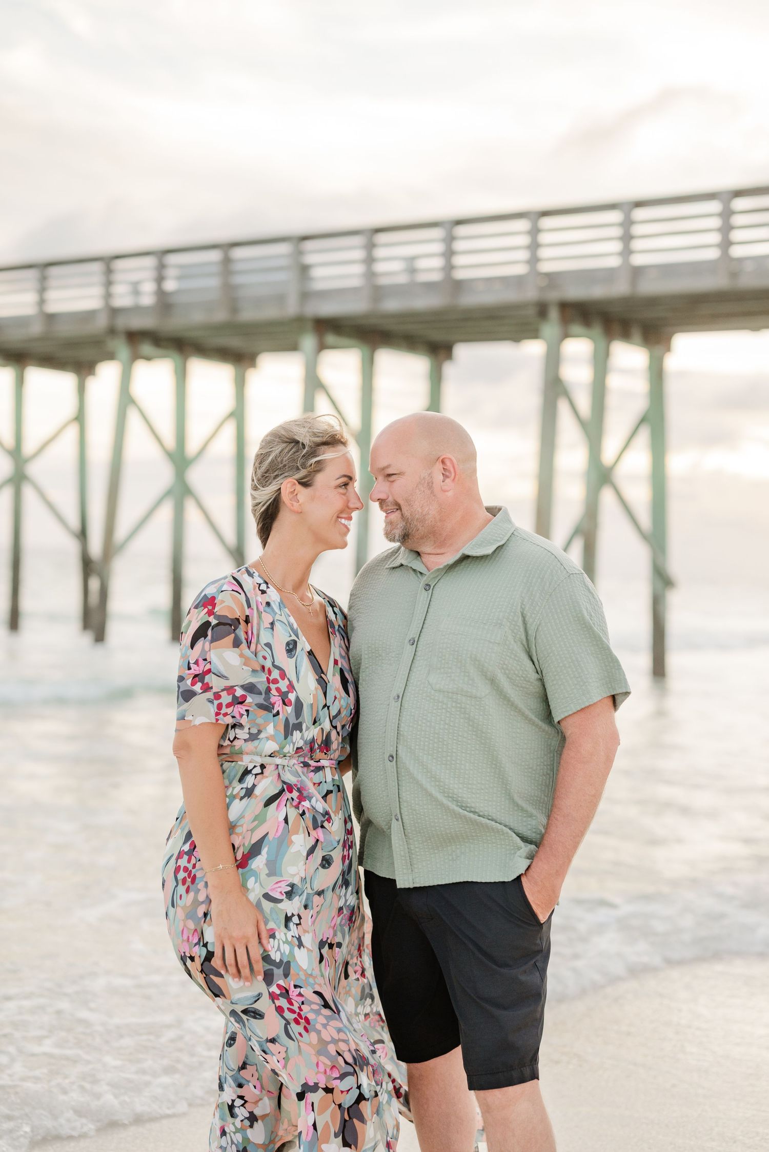 A couple poses near a wooden pier wearing summer attire during a beach photoshoot.