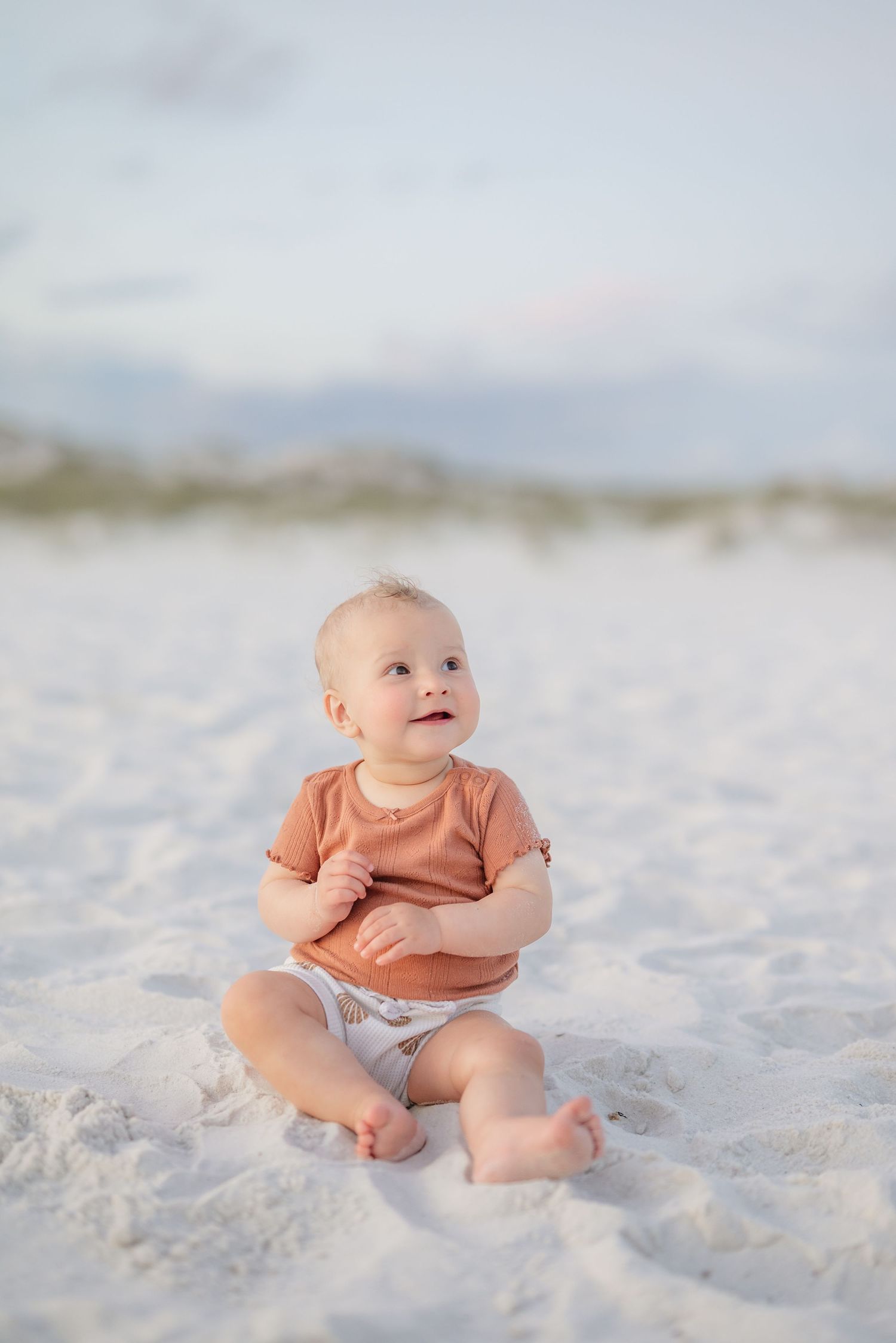 Baby wearing peachy colored shirt sitting and playing in white beach sand in Panama City Beach, Fl..