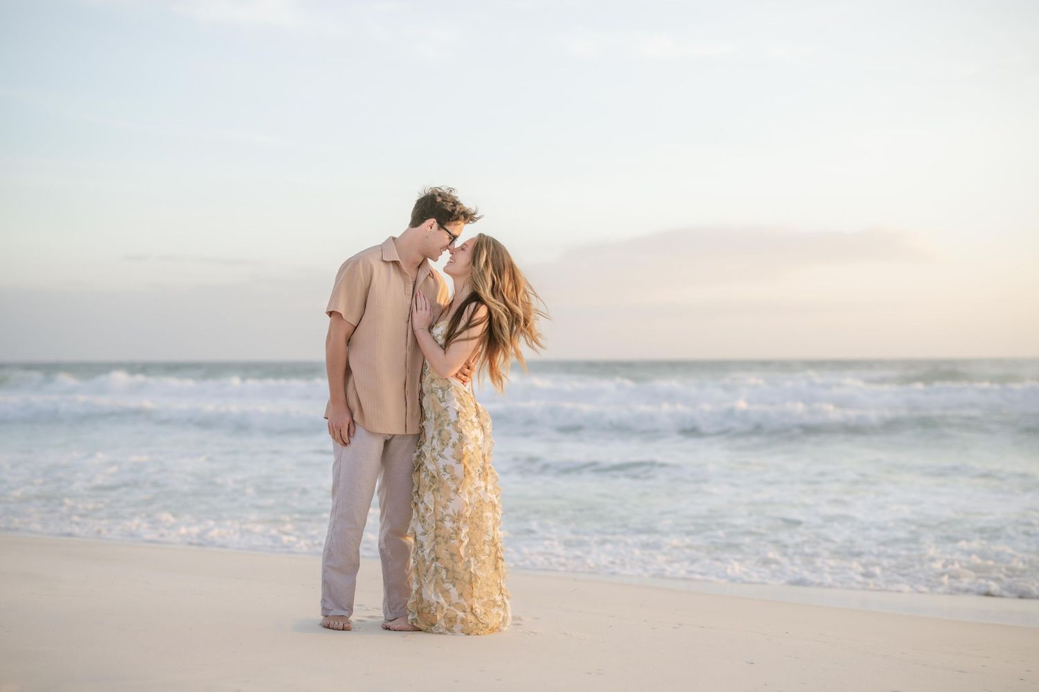 Couple sharing a romantic kiss on a sandy beach at sunset with gentle ocean waves in the background.
