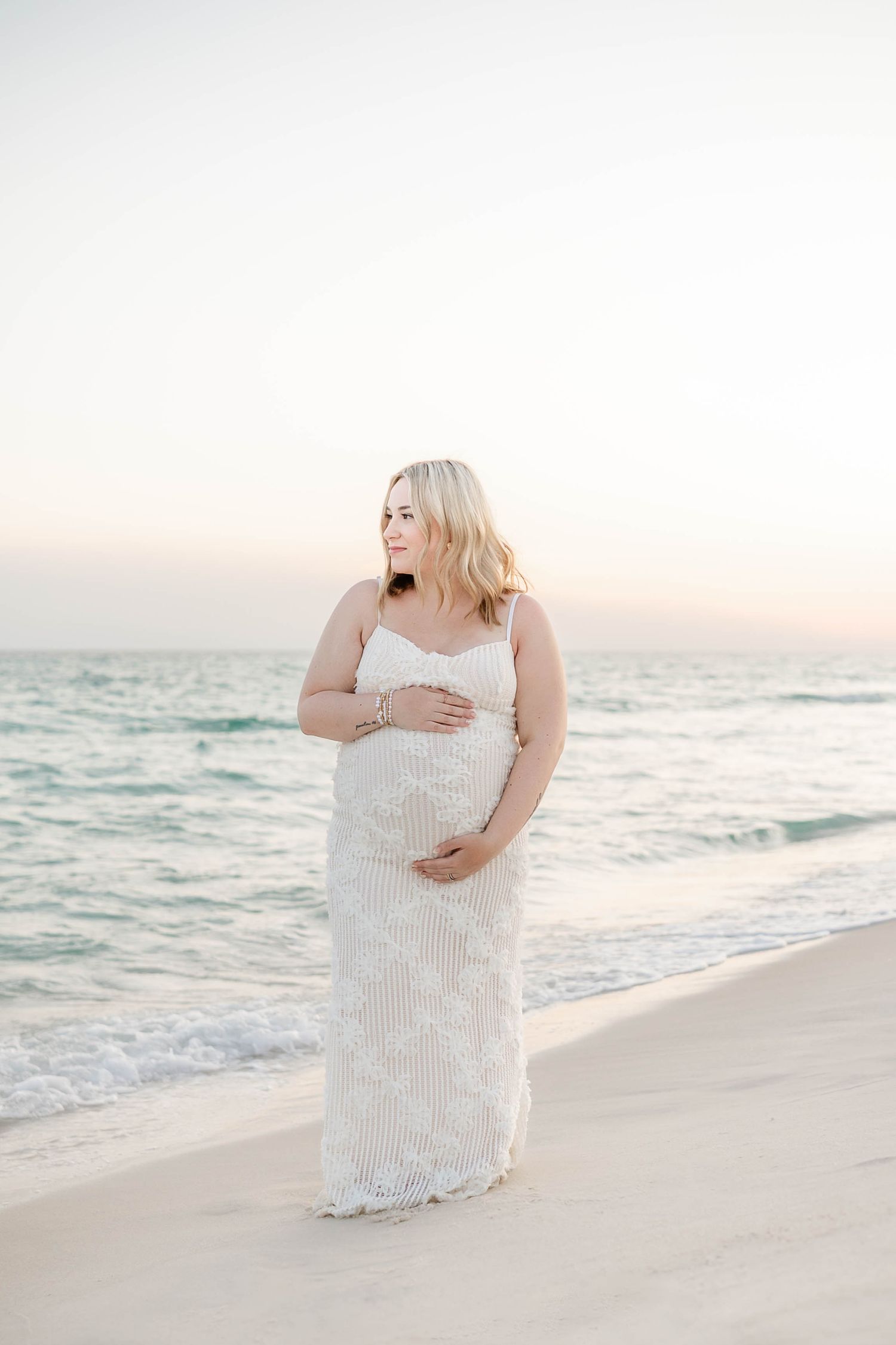 Woman in white dress standing on beach during sunset maternity photoshoot in Panama City Beach, Fl..