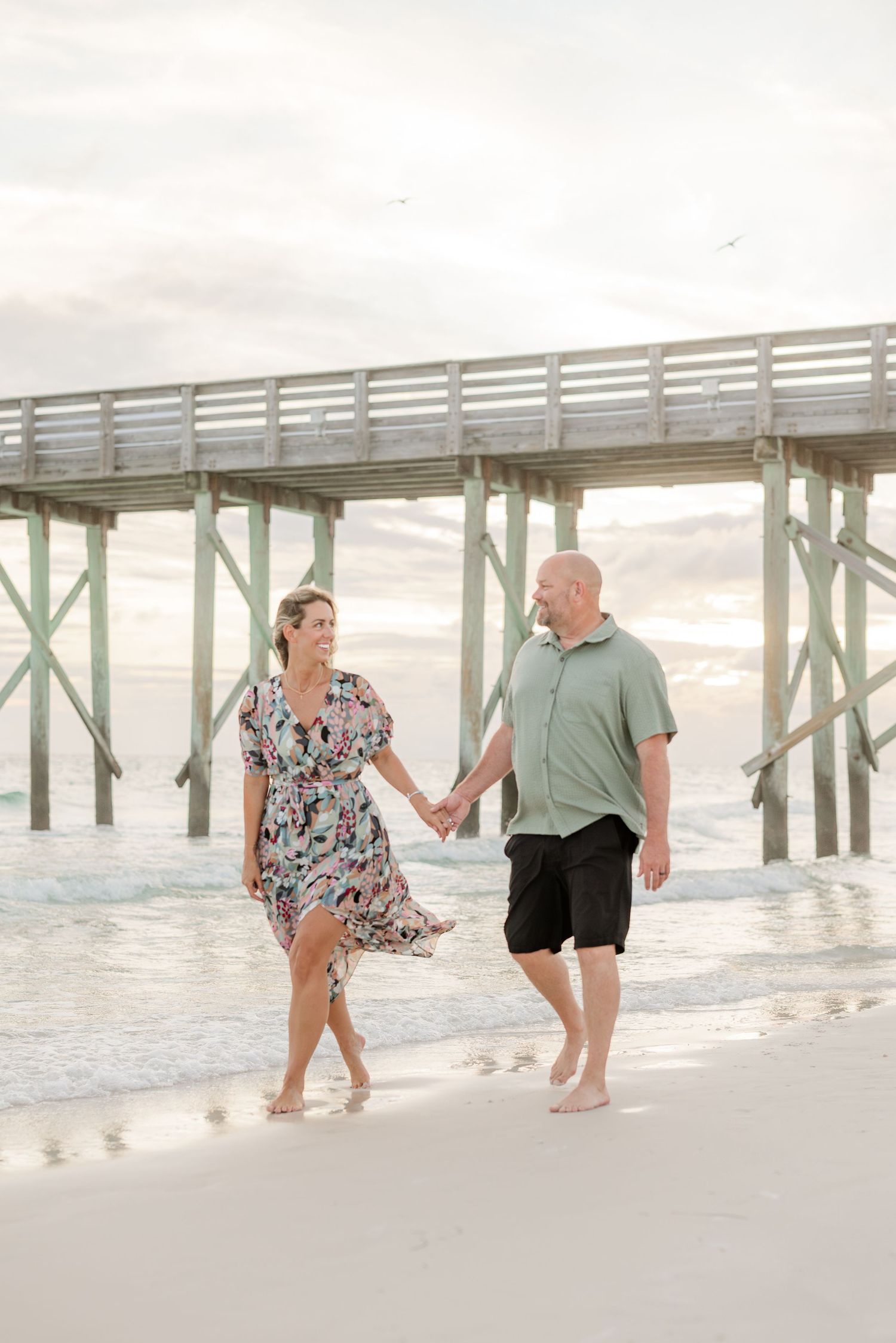 Couple walking playfully along a beach pier at sunset in casual summer attire.