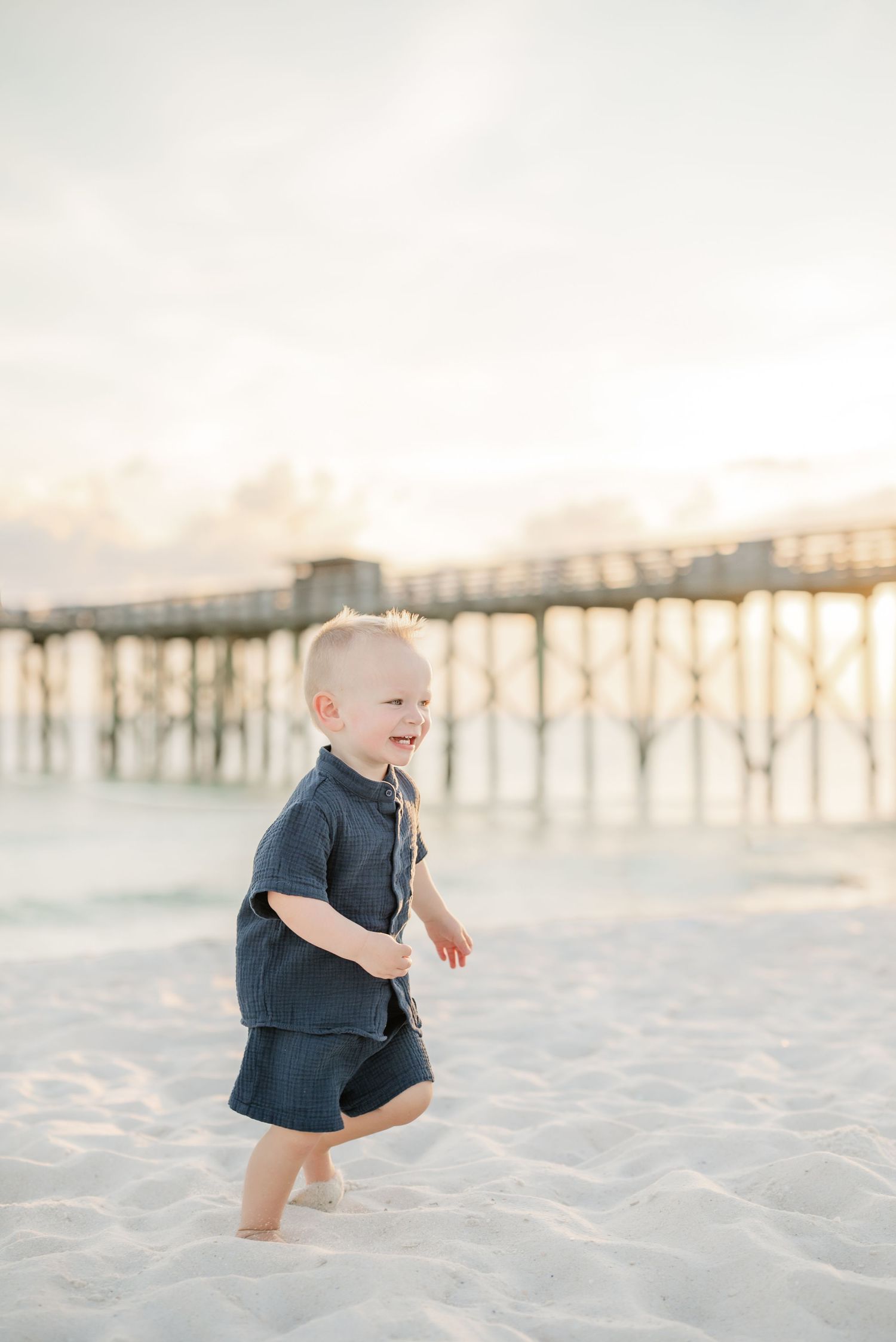 A child in navy outfit runs joyfully on beach sand with pier silhouetted in golden sunset.