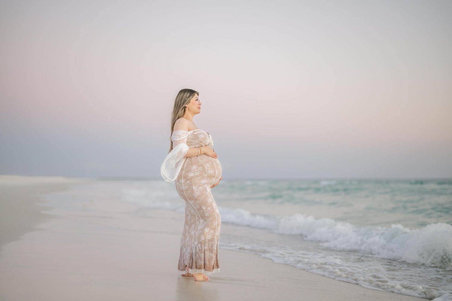 Pregnant woman in lace dress standing on beach at sunset, cradling baby bump with ocean waves behind her.