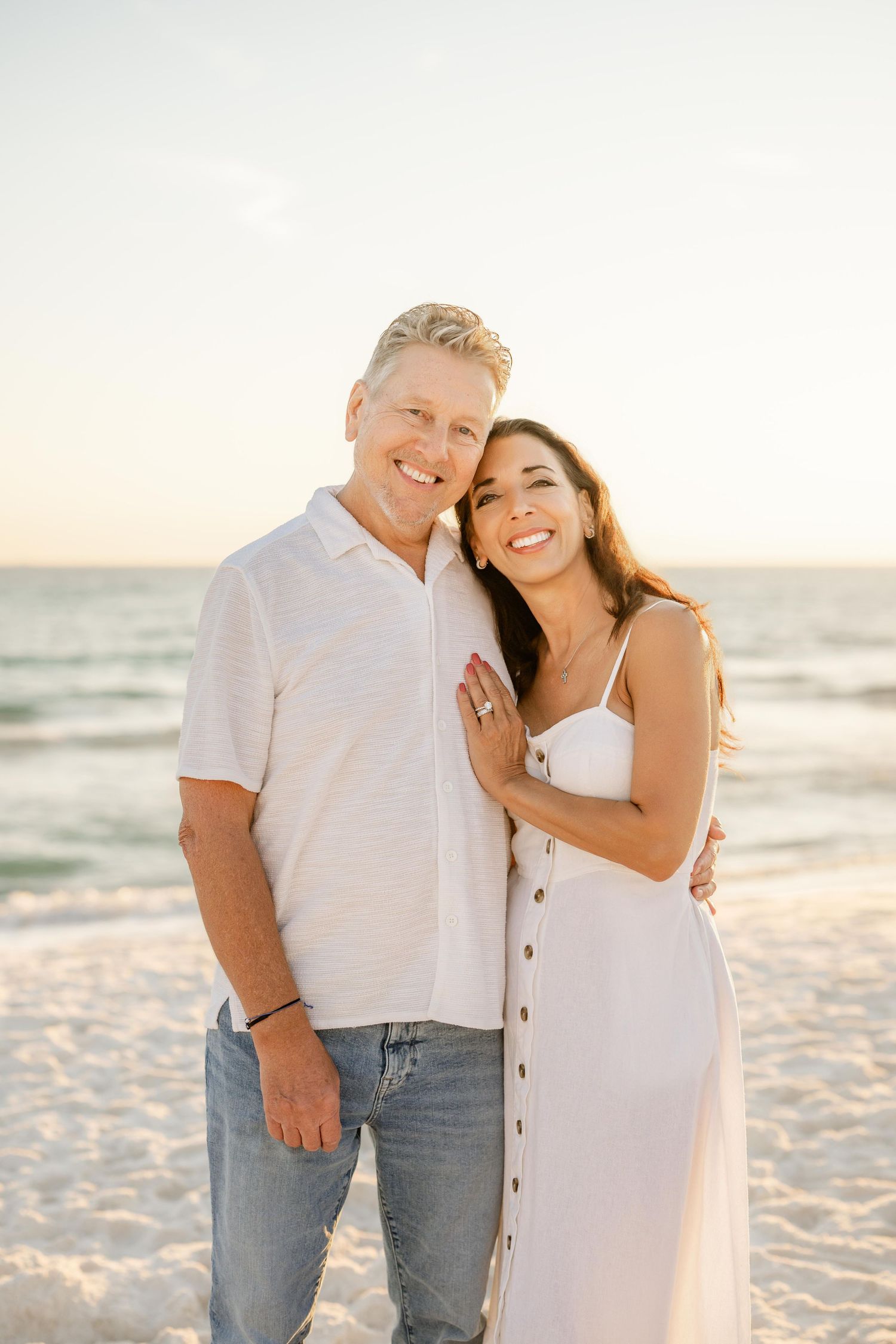Couple in white outfits sharing romantic moment on sandy beach at sunset.