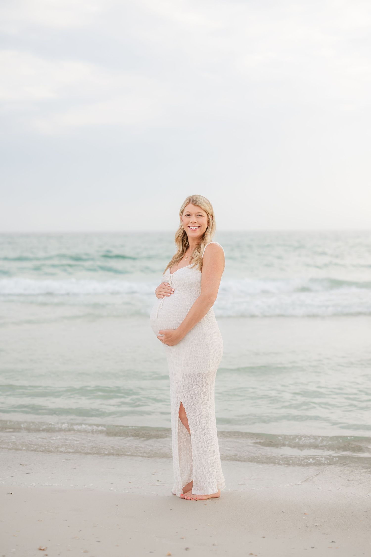 Pregnant woman in white dress posing on beach during golden hour photoshoot in Panama City Beach, Fl..