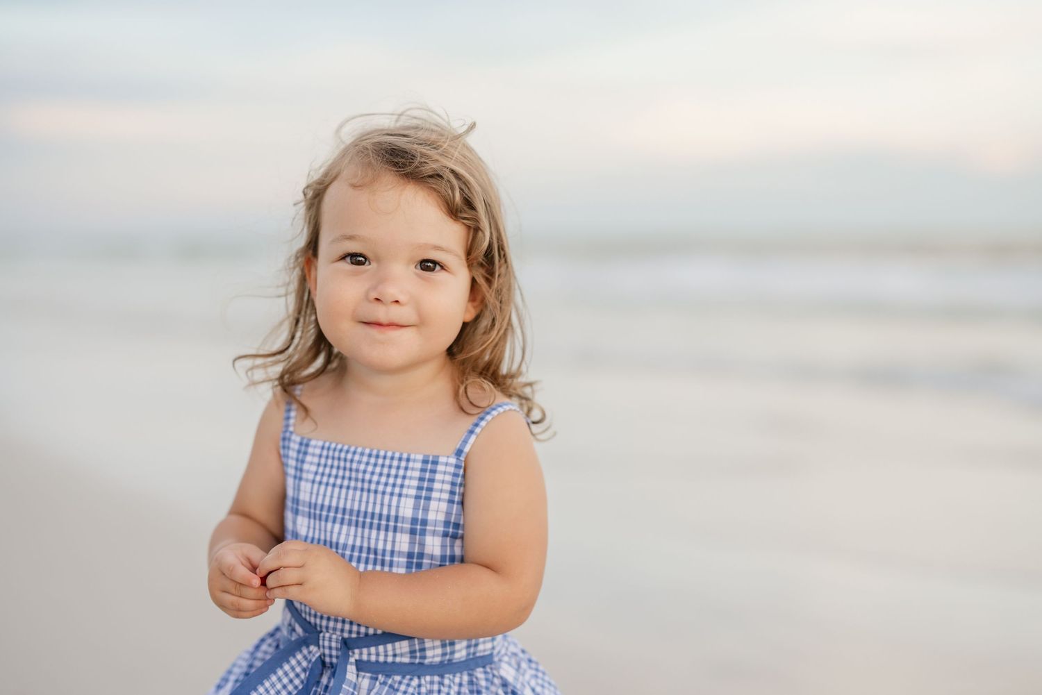 Child wearing blue gingham dress standing on sandy beach during sunset.