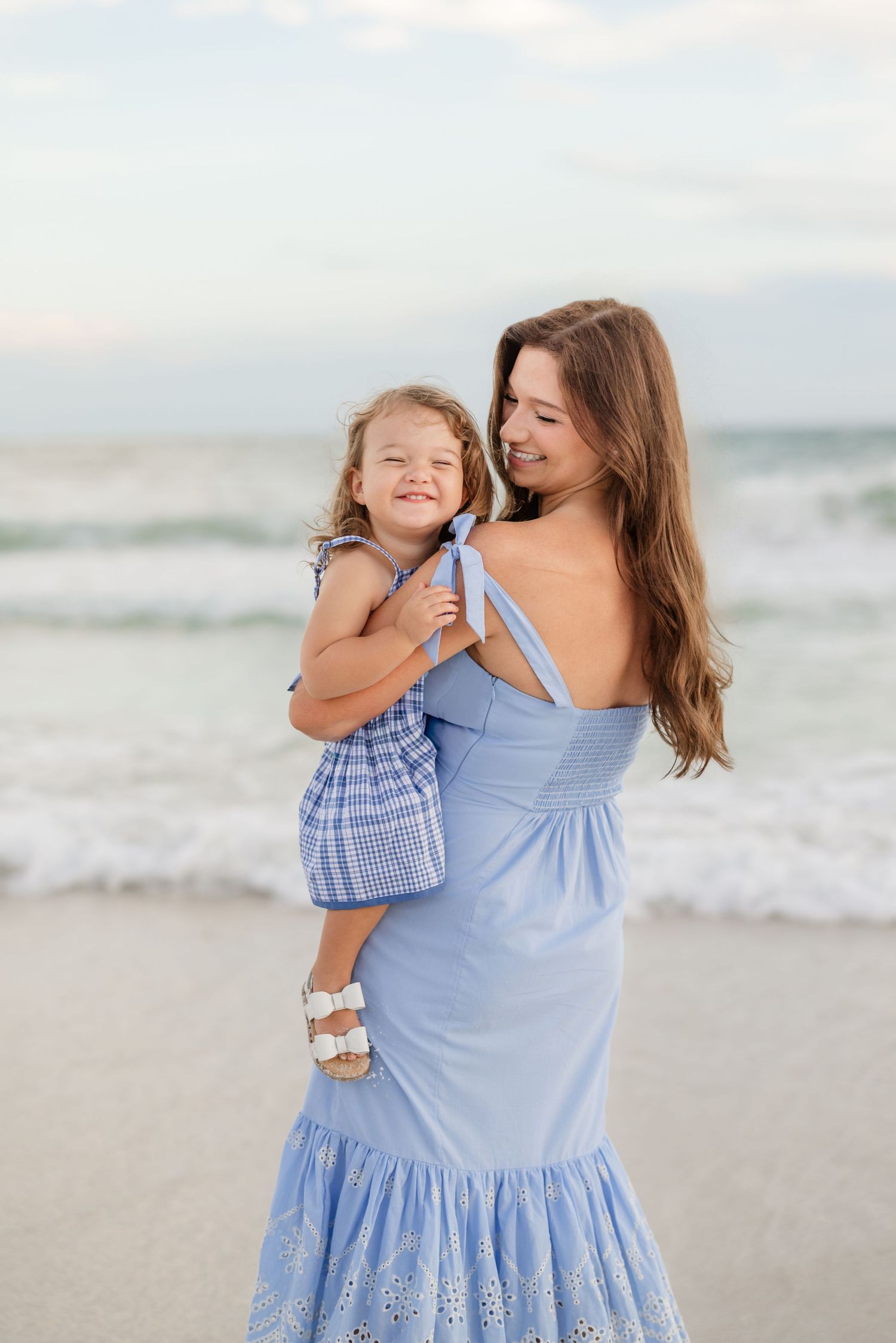 Mother and daughter in coordinating blue outfits embrace on a sandy beach at sunset.