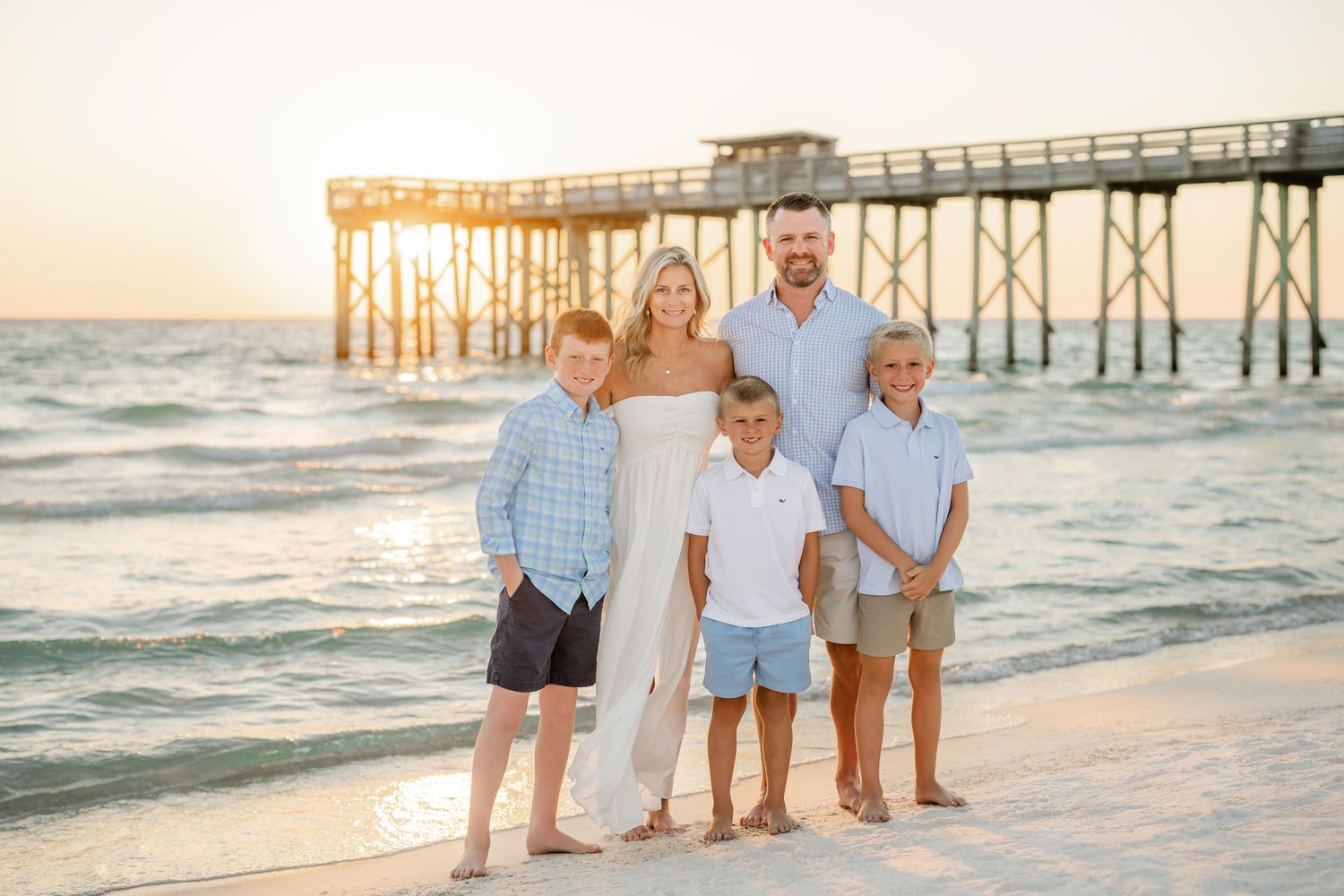 A family poses together on a beach at sunset with a wooden pier in the background.