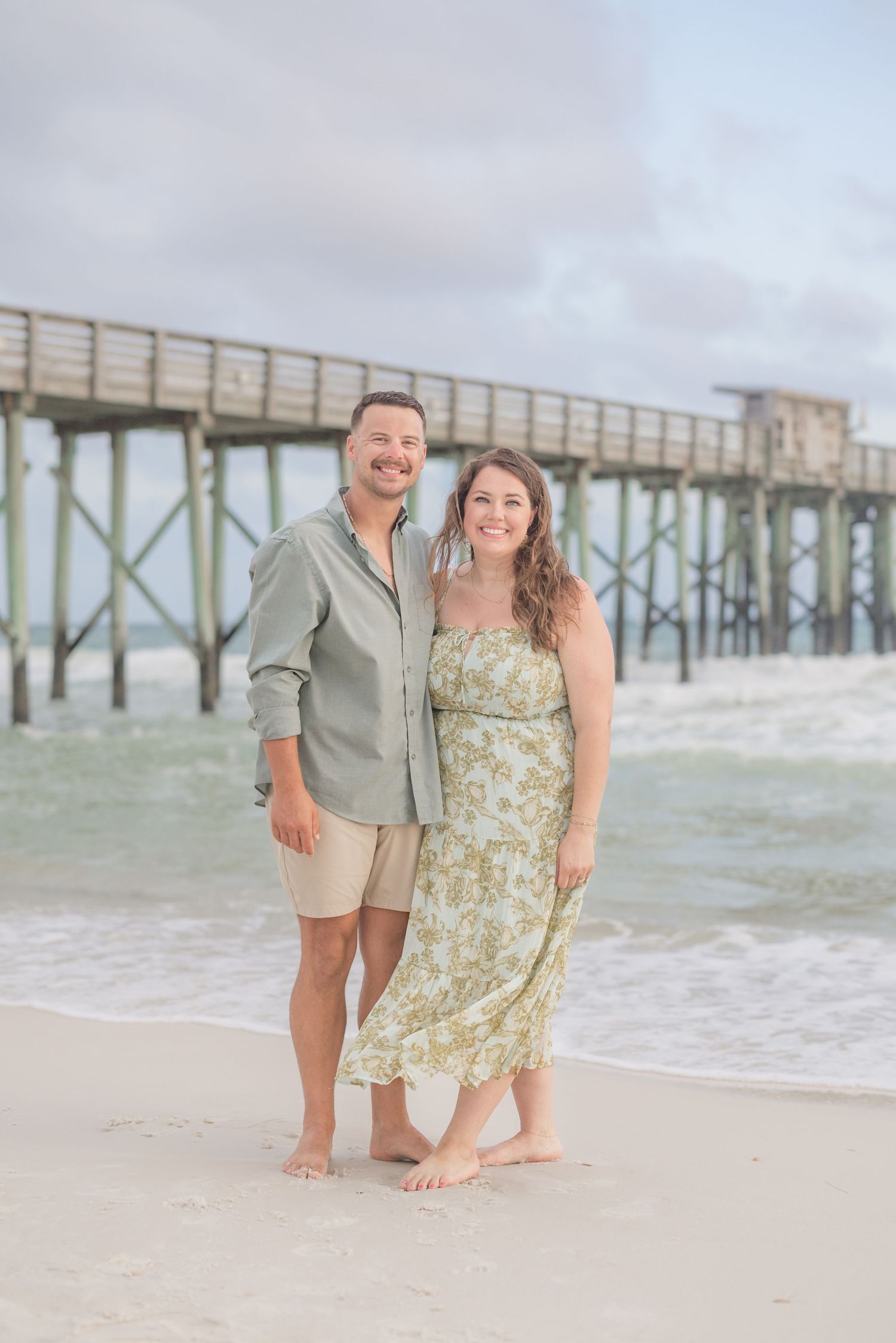 Couple posing playfully on beach with wooden pier and ocean waves in background.