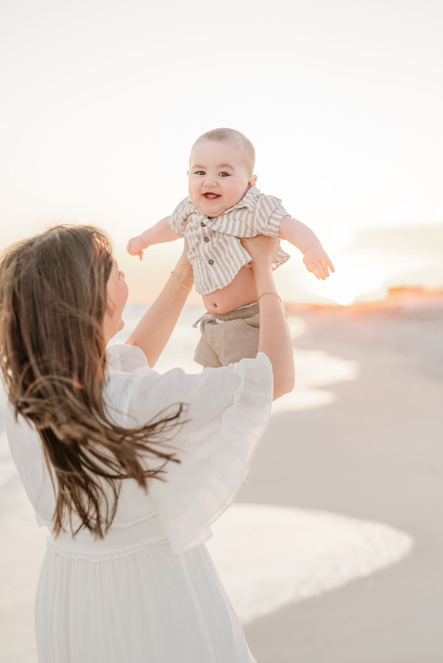 Parent lifting and playing with a smiling baby against a bright sunset backdrop.