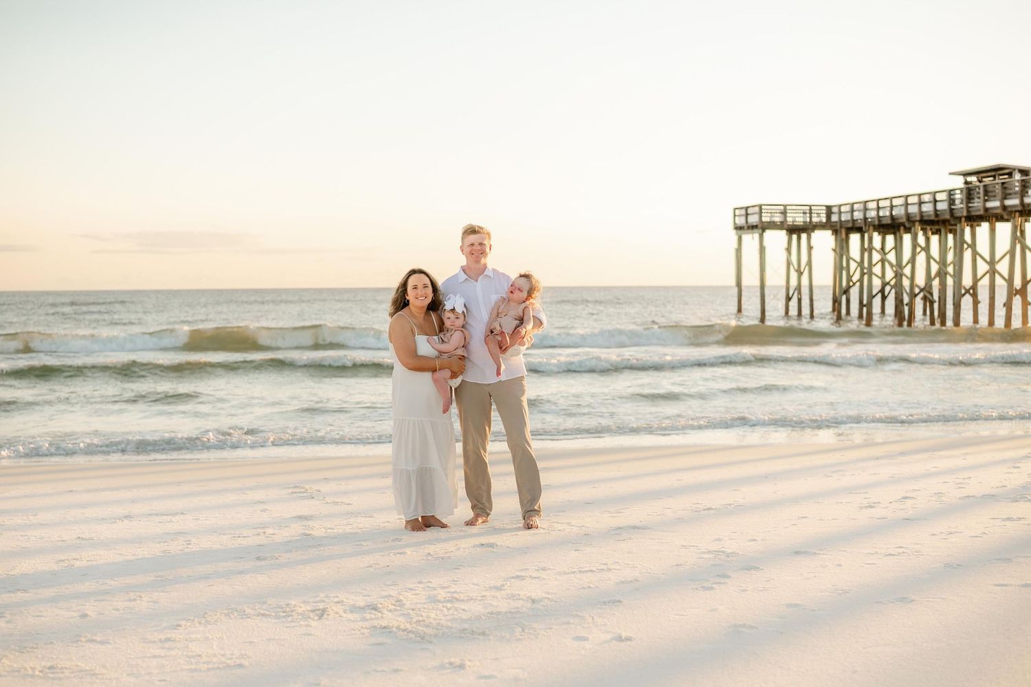 Casual family beach portraits with wooden pier and ocean waves at sunset.