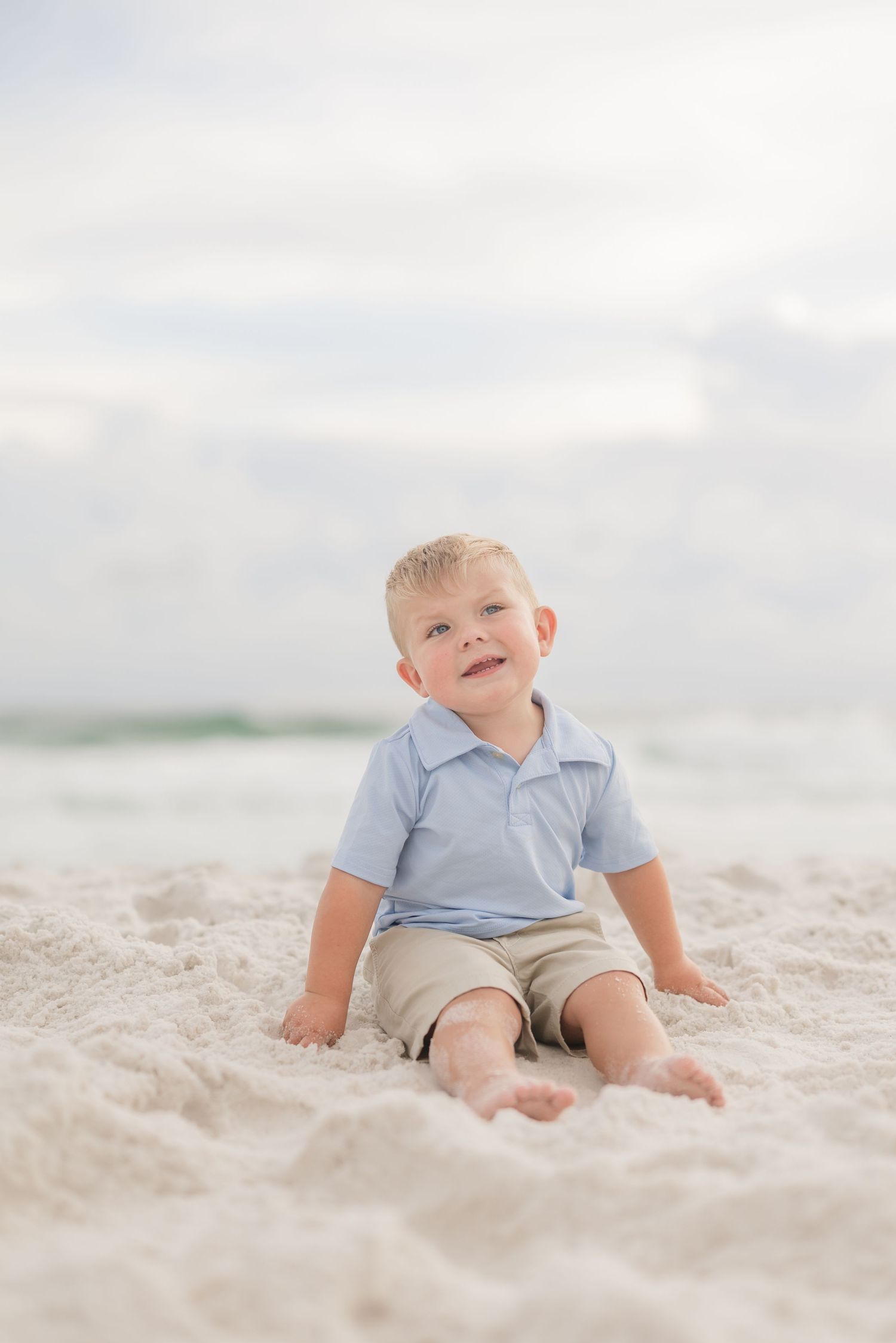 Young child sitting on white sand beach looking up with joyful expression in Panama City Beach, Fl..