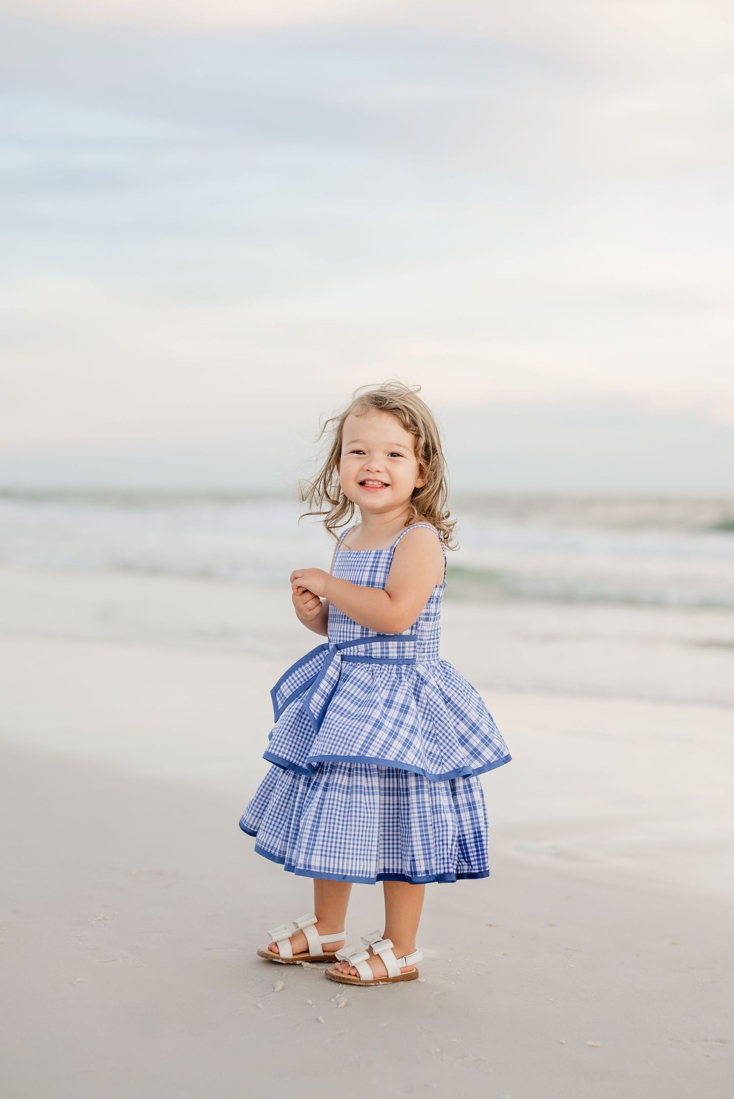 A child in a blue gingham dress twirls and plays on a misty beach at sunset.